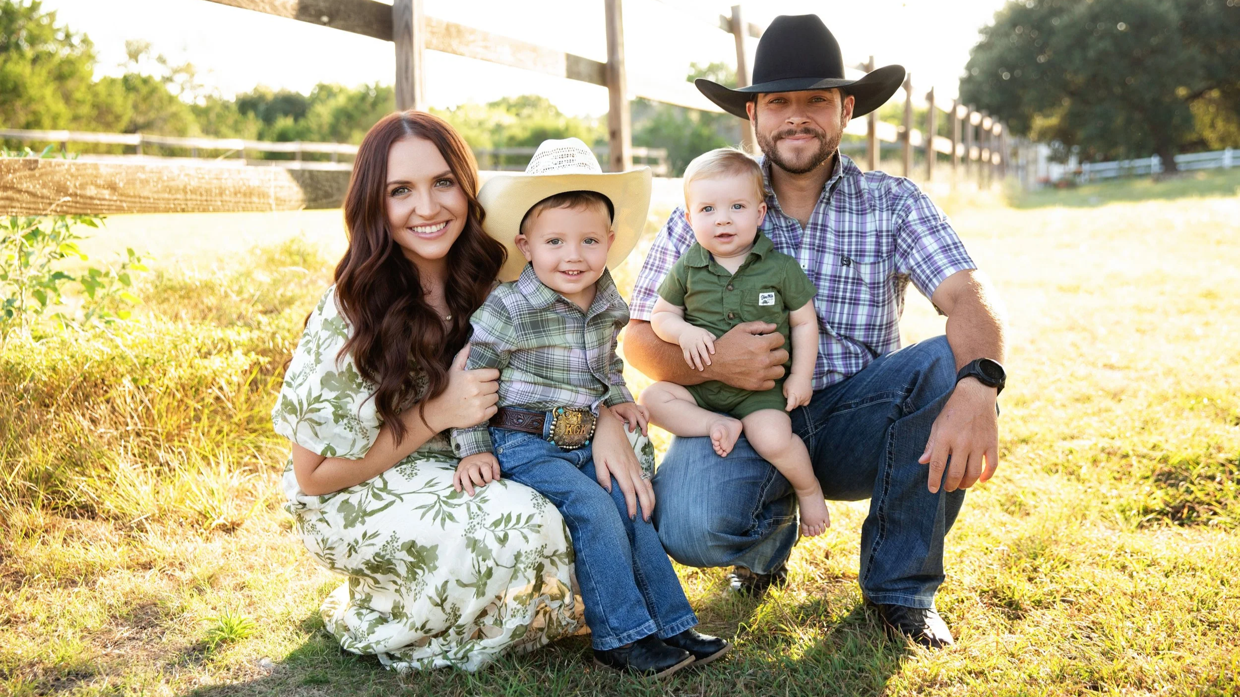 A family of five sitting outdoors in a rural setting, smiling and dressed in country-style clothing, with a wooden fence and trees in the background.