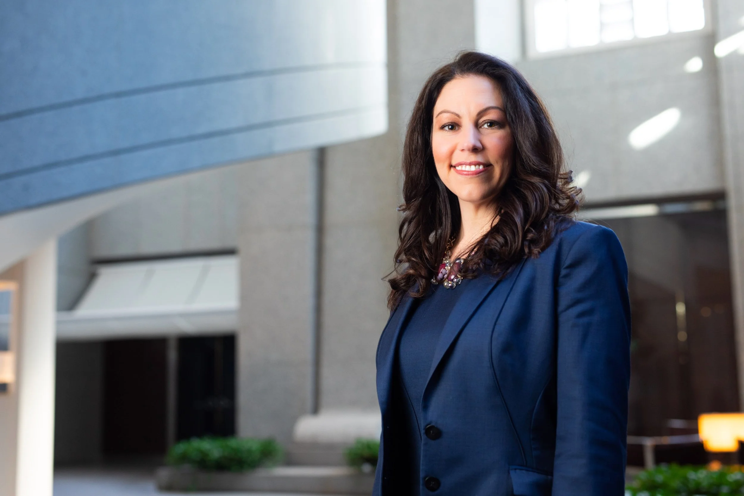 A woman with long dark hair wearing a navy blue blazer and a patterned necklace, smiling, standing in a modern building with large windows and a bright interior.