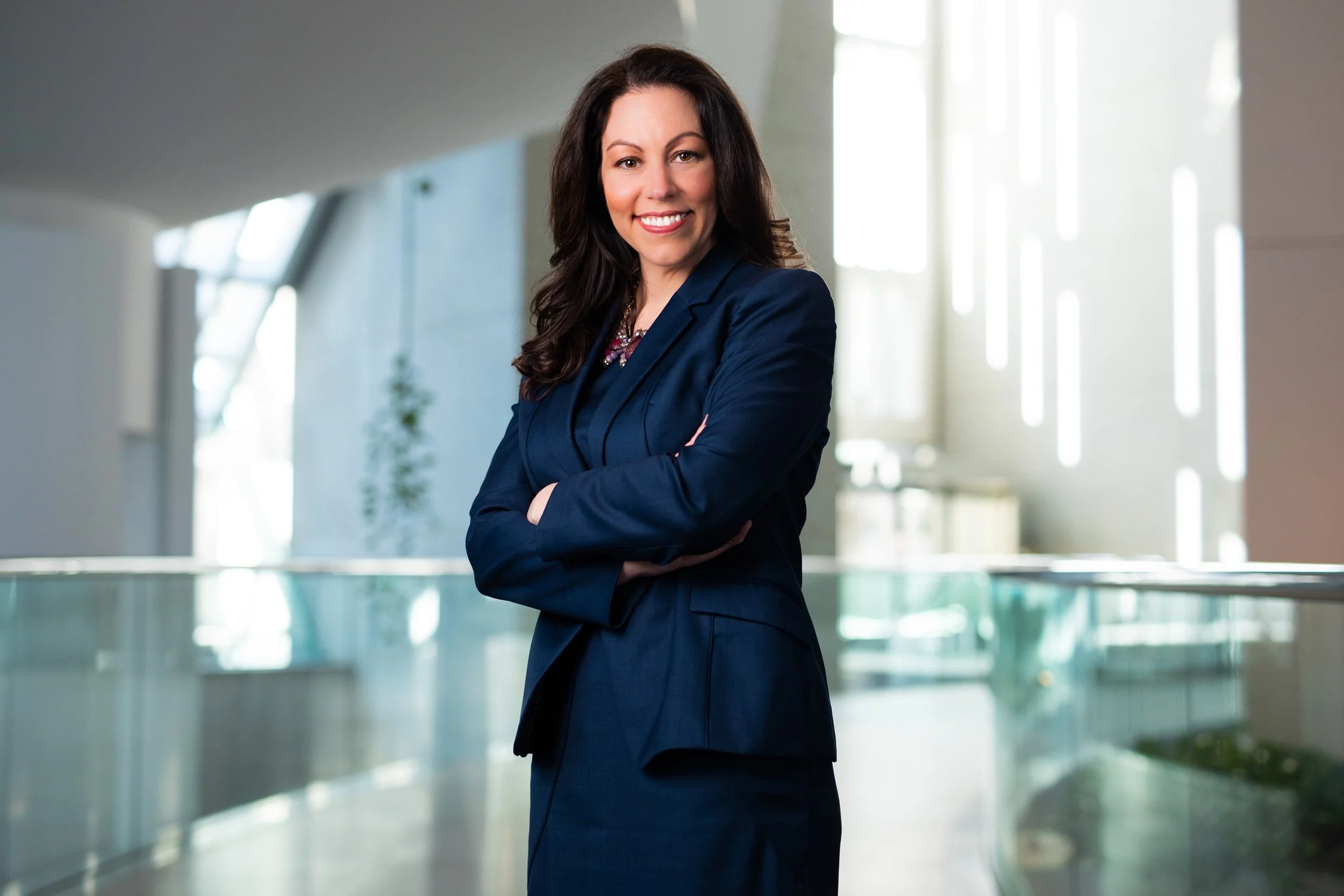 A professional woman in a dark blue suit smiling with arms crossed in an office building with modern architecture and large windows.