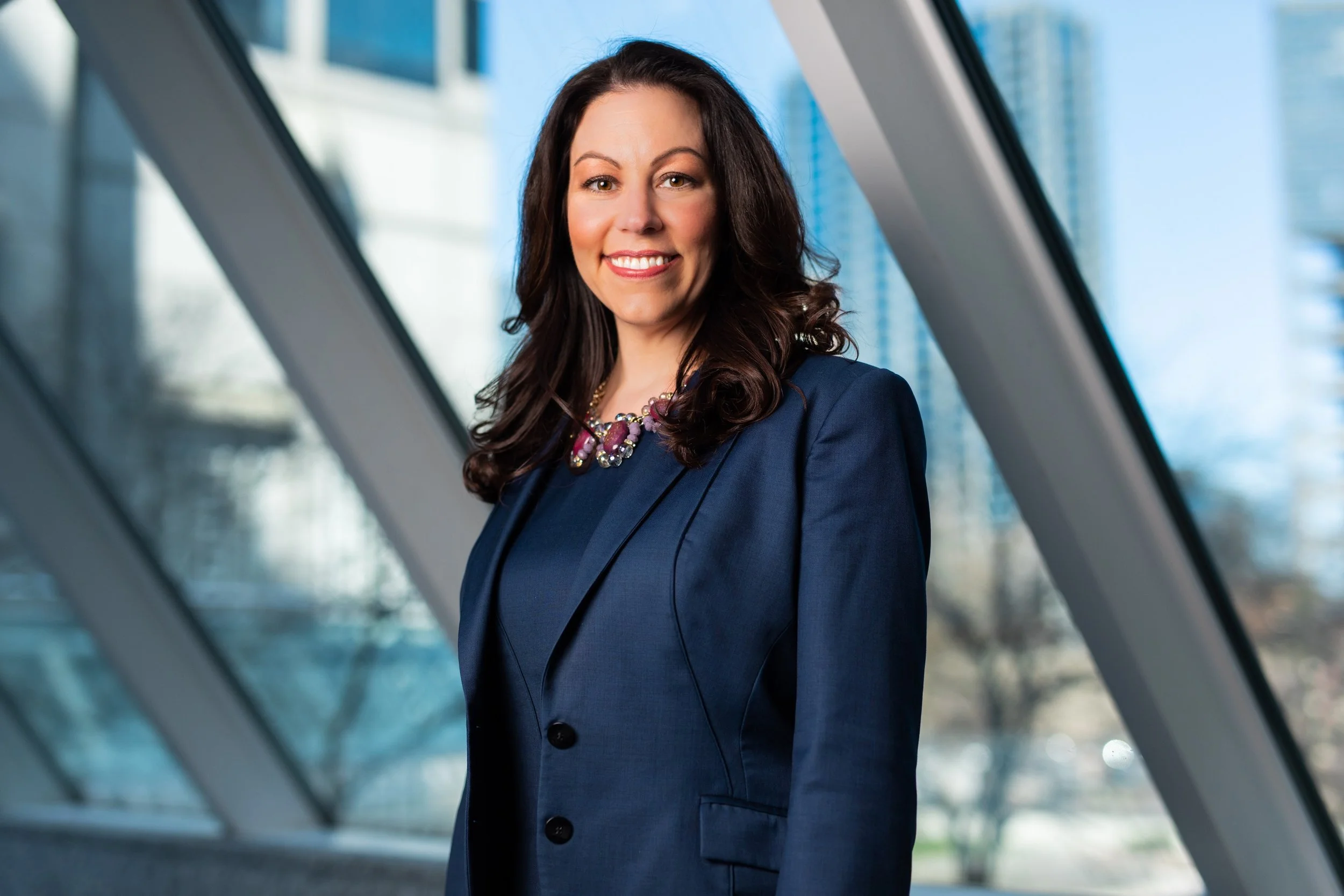 Portrait of a smiling woman with dark hair wearing a navy blazer and a chunky necklace, standing in front of a modern glass building with a blue sky in the background.