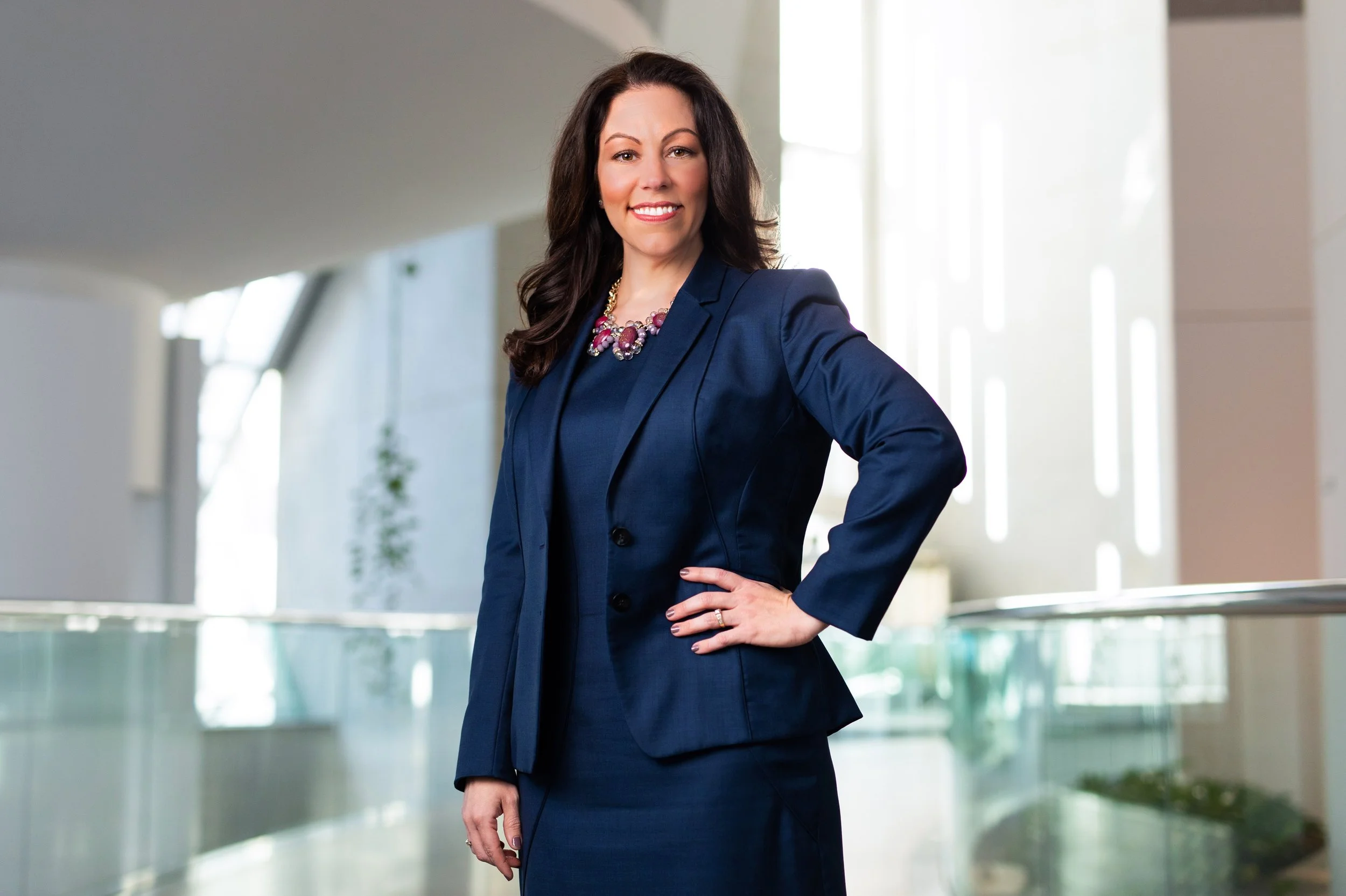 A confident woman with dark brown hair, wearing a navy blue business suit and a statement necklace, standing indoors near large windows with a modern architectural background.