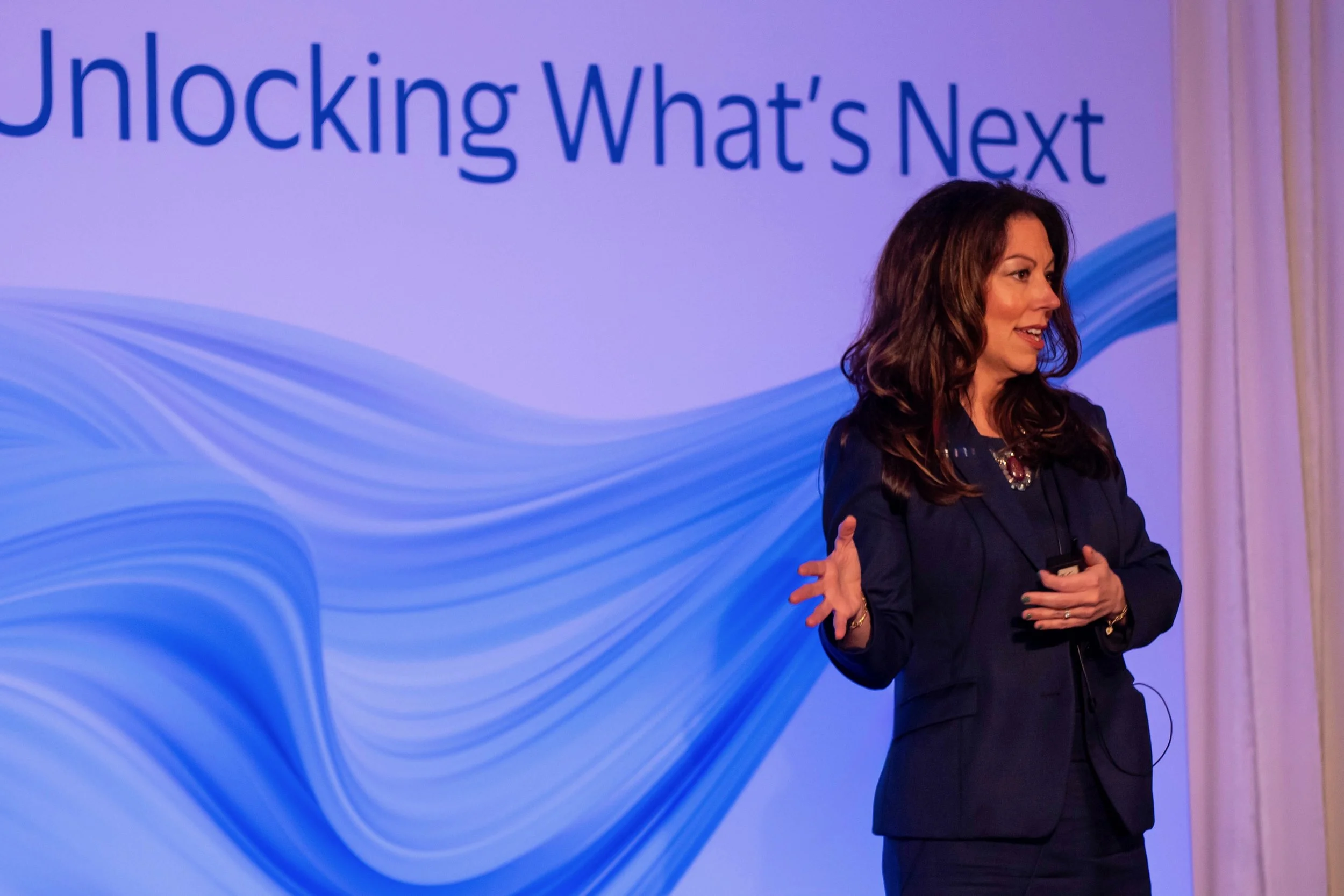 A woman speaking at a conference or event, standing in front of a purple and blue background with the words 'Unlocking What's Next' written on it.