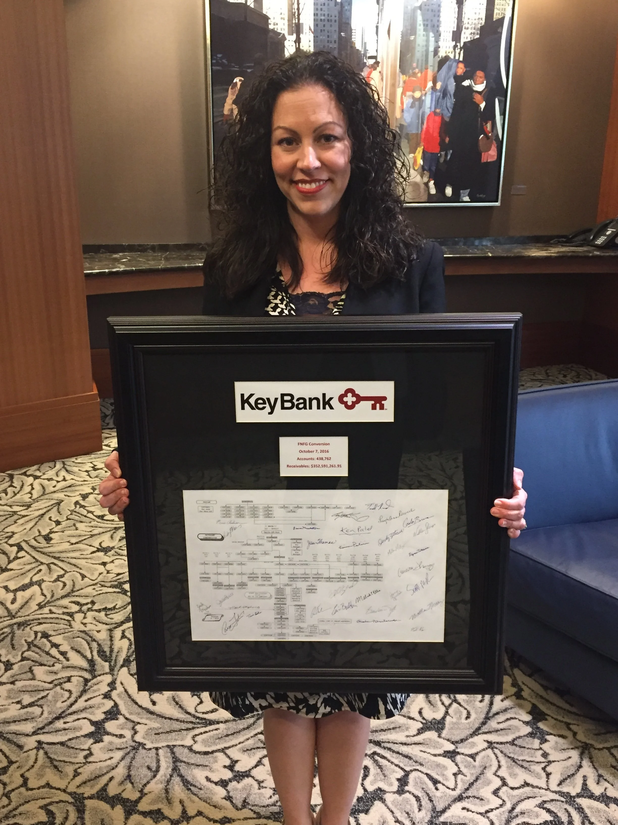 A woman with curly hair holding a framed signed document from KeyBank. She is smiling and standing in front of a painting and a brown wall. The event took place on October 7, 2016.