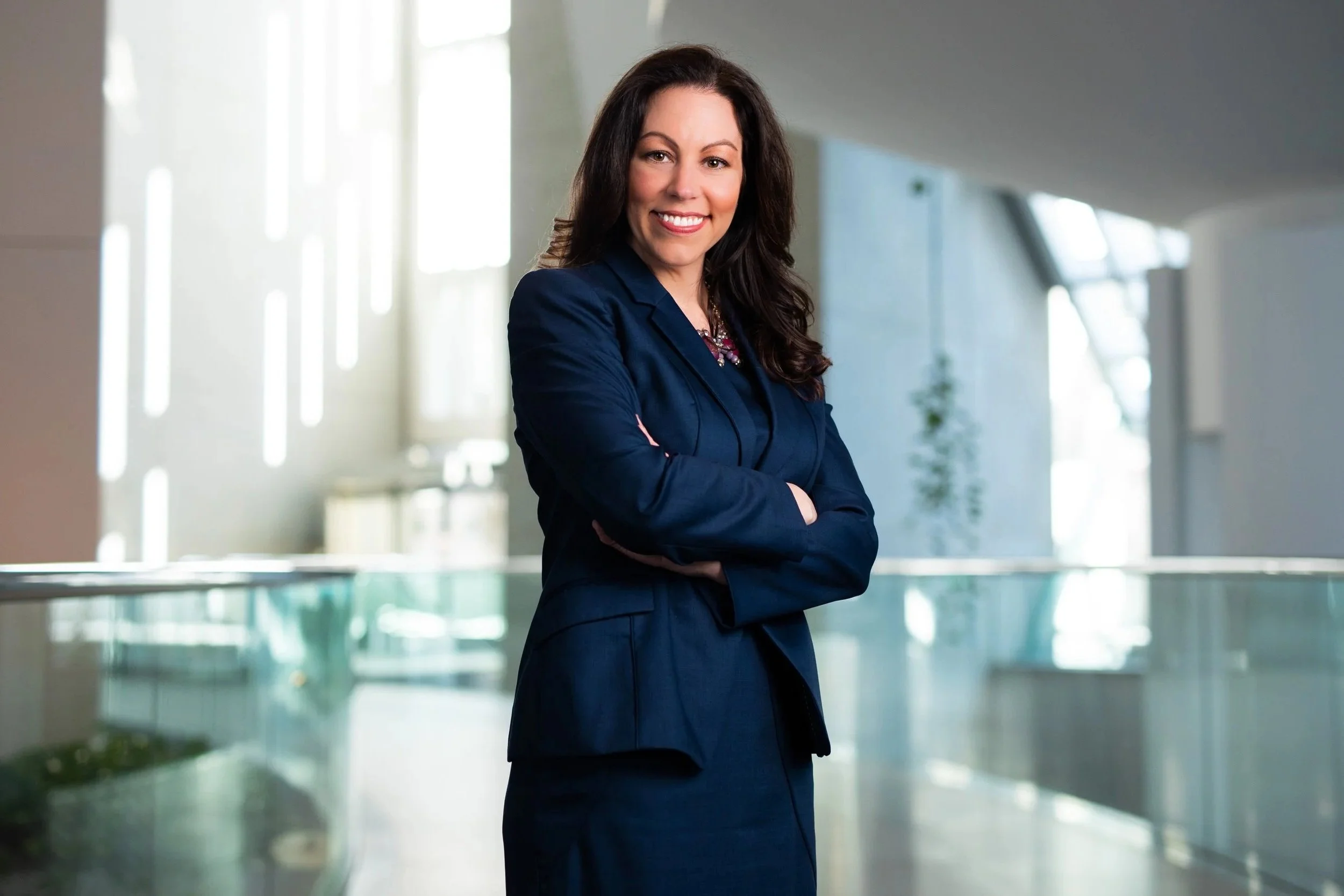 A woman with long dark hair, wearing a navy blazer and a patterned blouse, is smiling and standing with her arms crossed in an airy, modern office building with large windows.