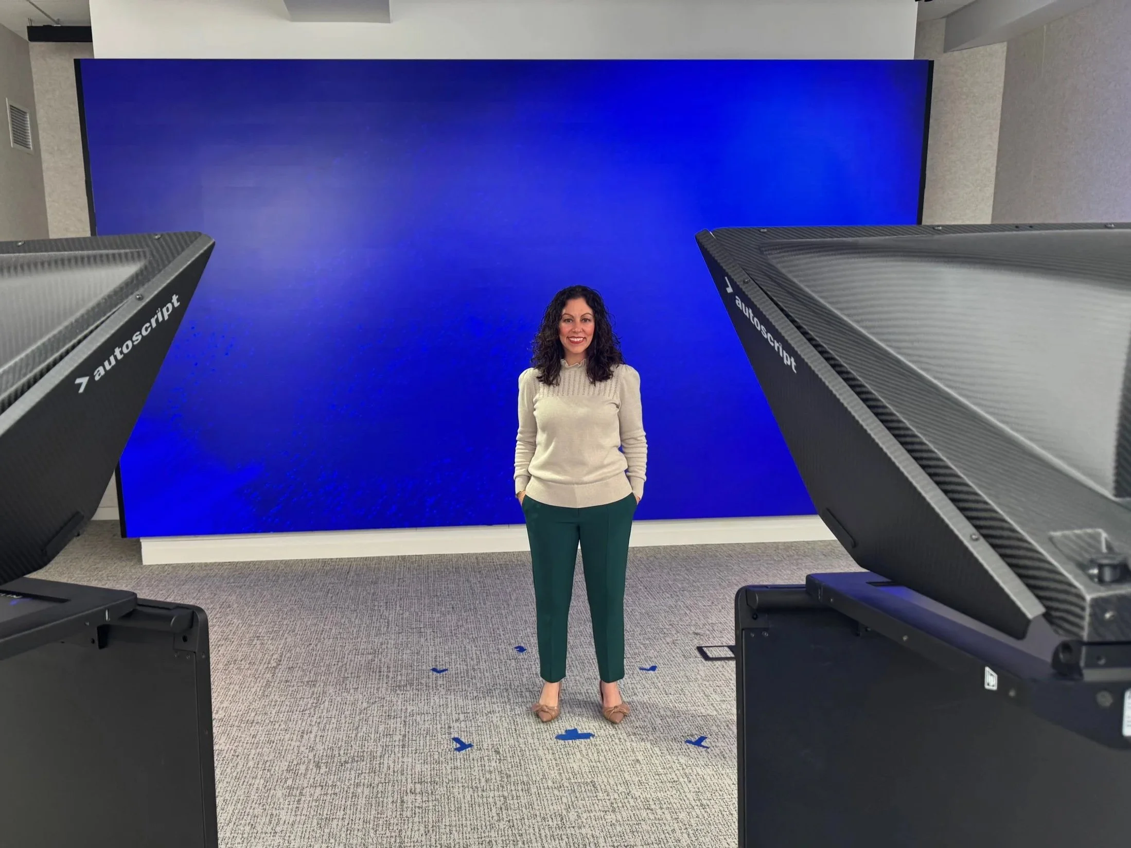A woman in a beige sweater and green pants standing in front of a large blue screen inside a room, with large floodlights labeled 'autoscript' on either side.