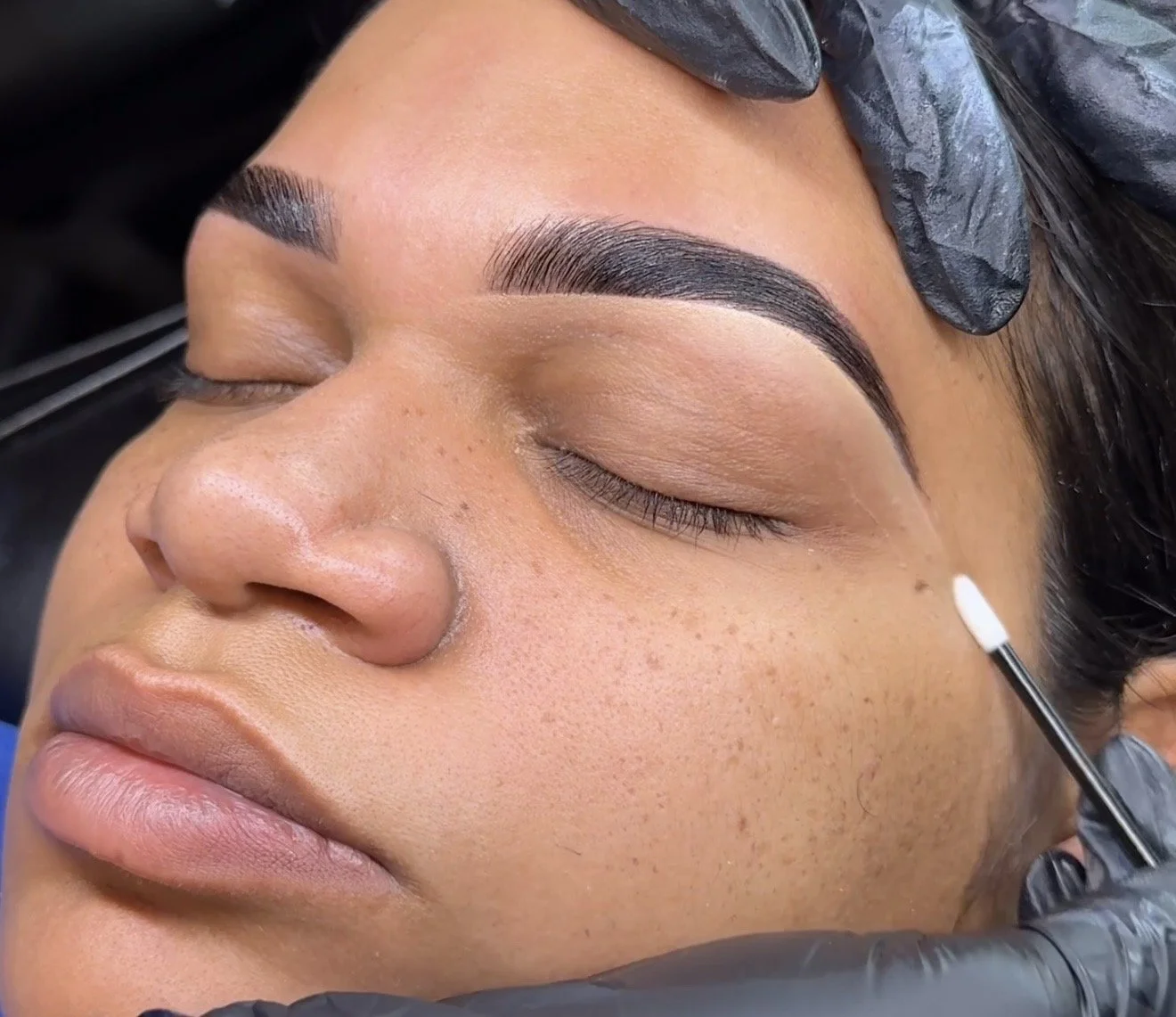 Close-up of a woman with freshly done eyebrow microblading, lying with eyes closed, a gloved technician using a small brush near her brow in a beauty salon.