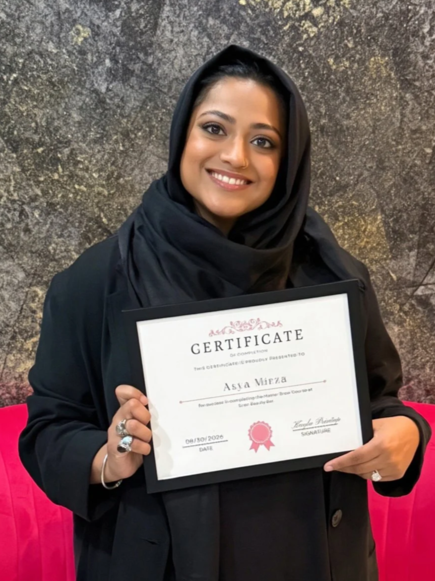 Woman in a black hijab holding a certificate smiling, standing in front of a textured wall and a pink cushion.