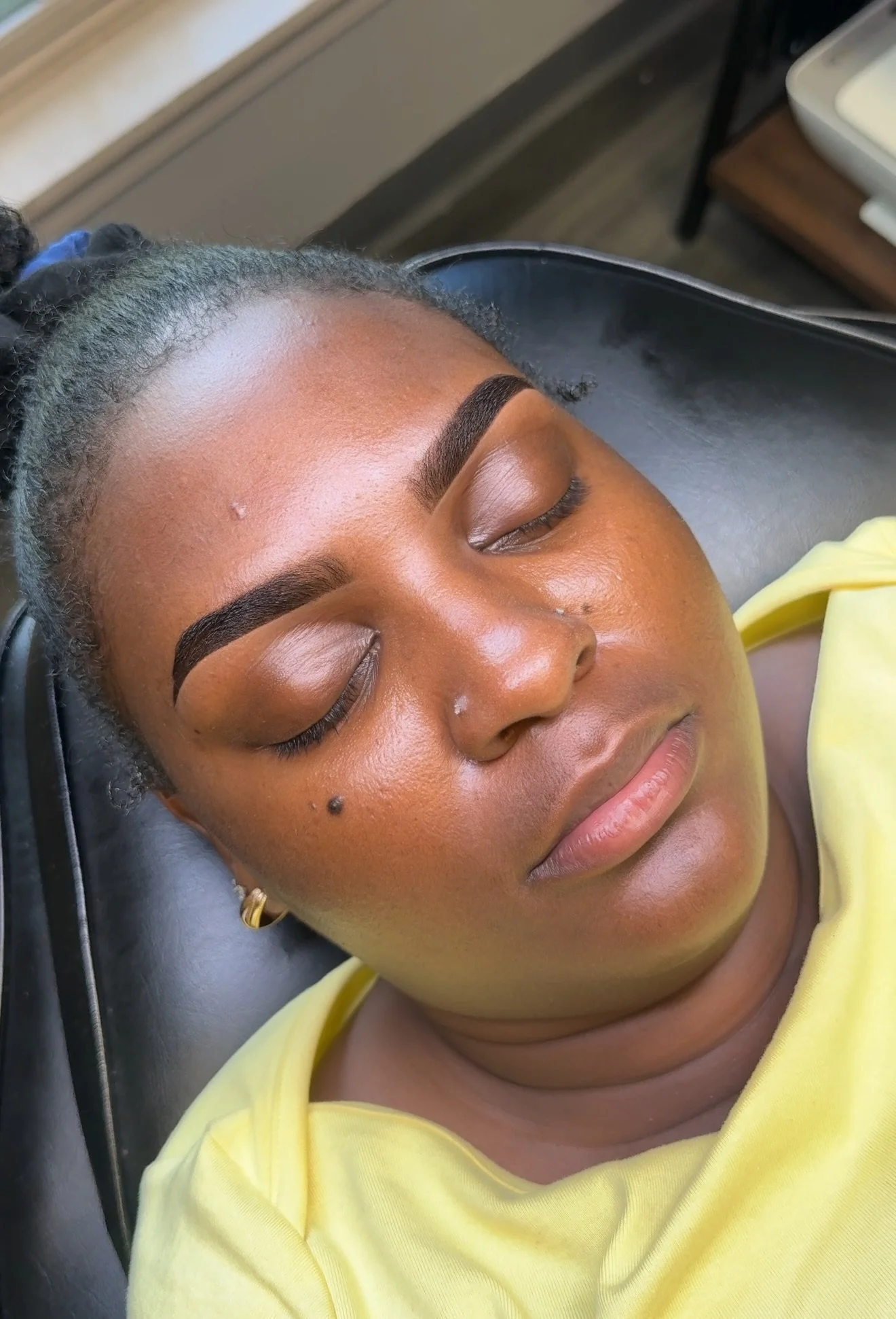 Close-up of a woman with well-groomed eyebrows, lying down with eyes closed, wearing a yellow top and earrings, possibly at a spa or salon, with a dark chair or bed beneath her head.