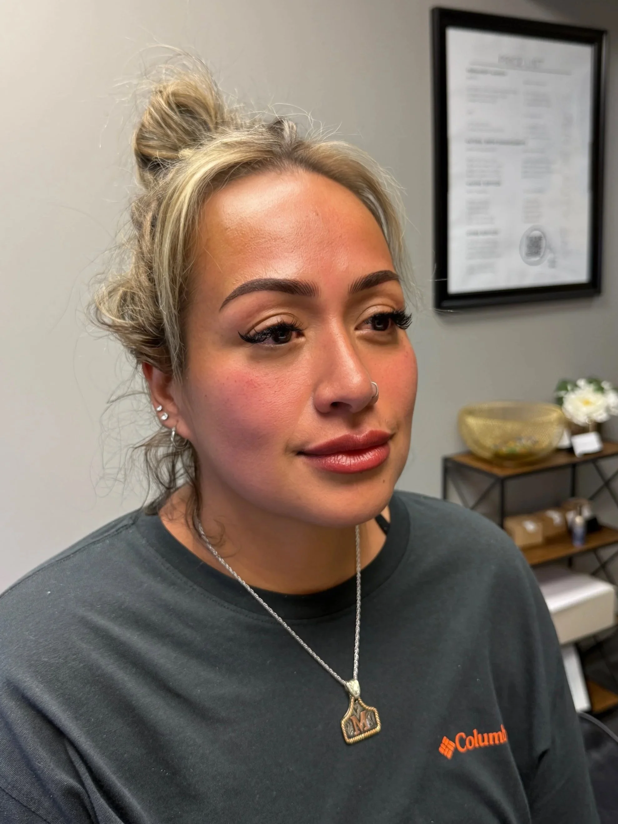 Close-up of a woman with blonde hair styled in a messy bun, wearing a dark gray Columbia sweatshirt and a necklace with a pendant, standing indoors with blurred decor and a framed document on the wall in the background.