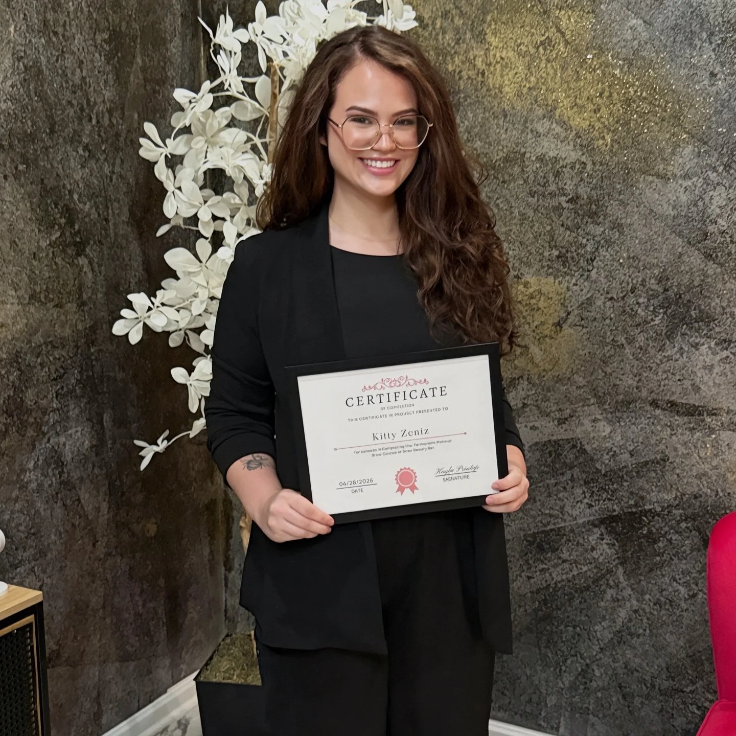 A young woman with long brown curly hair and glasses, dressed in black, smiling and holding a framed certificate of completion in front of a decorative white floral arrangement and dark textured wall.