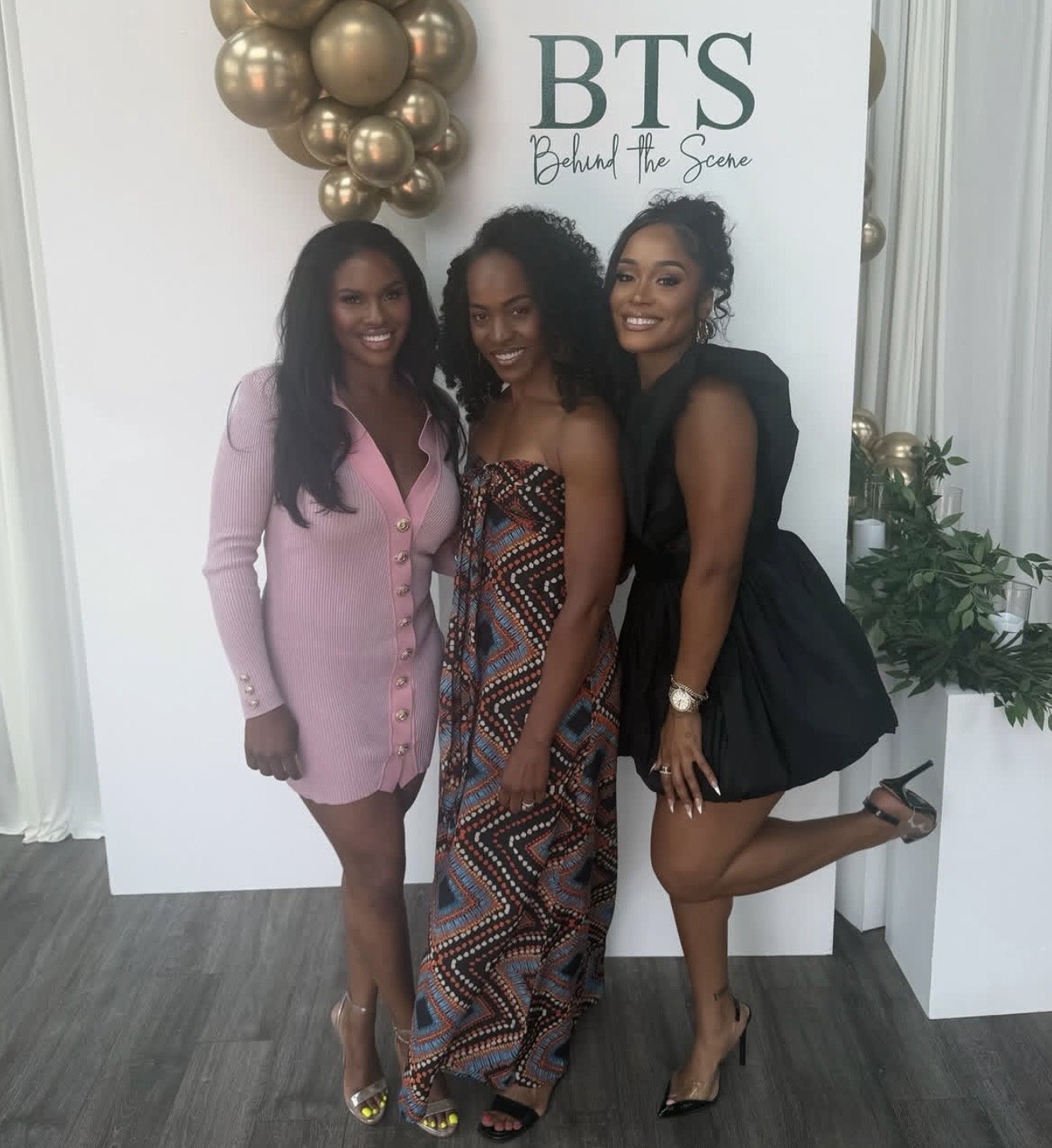 Three women posing together at an event with a backdrop that reads 'BTS Behind the Scene.' They are dressed fashionably, with one woman in a pink dress, another in a patterned gown, and the third in a black dress. They are smiling and appear joyful.