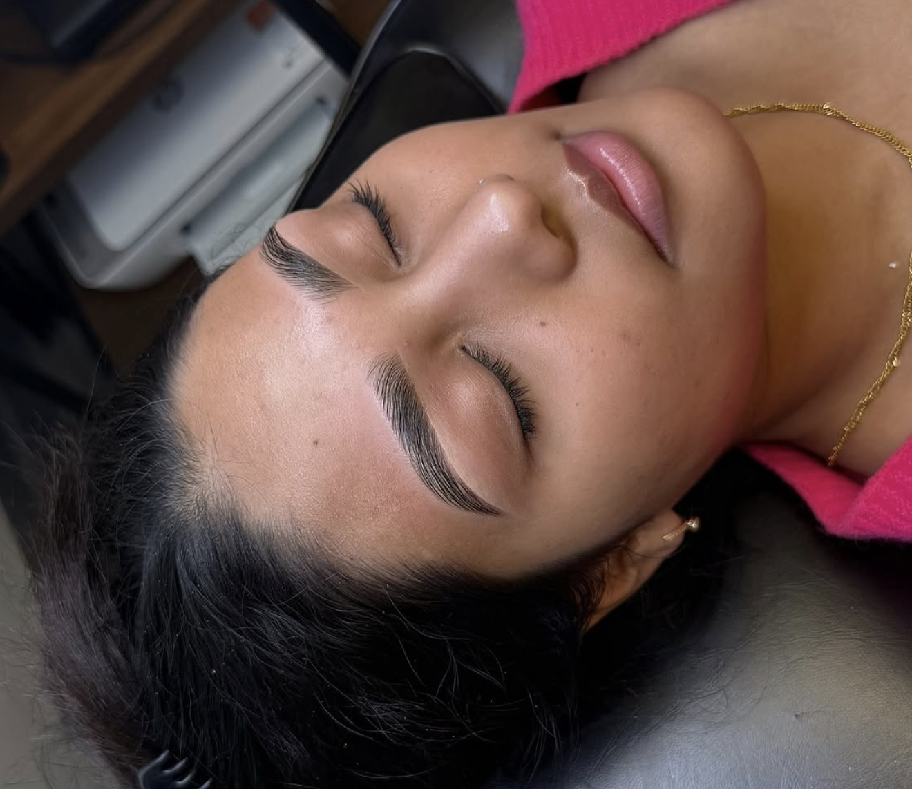 Close-up of a woman with freshly done eyelash extensions, lying on a black surface, with her eyes closed, wearing a pink top and a gold necklace.