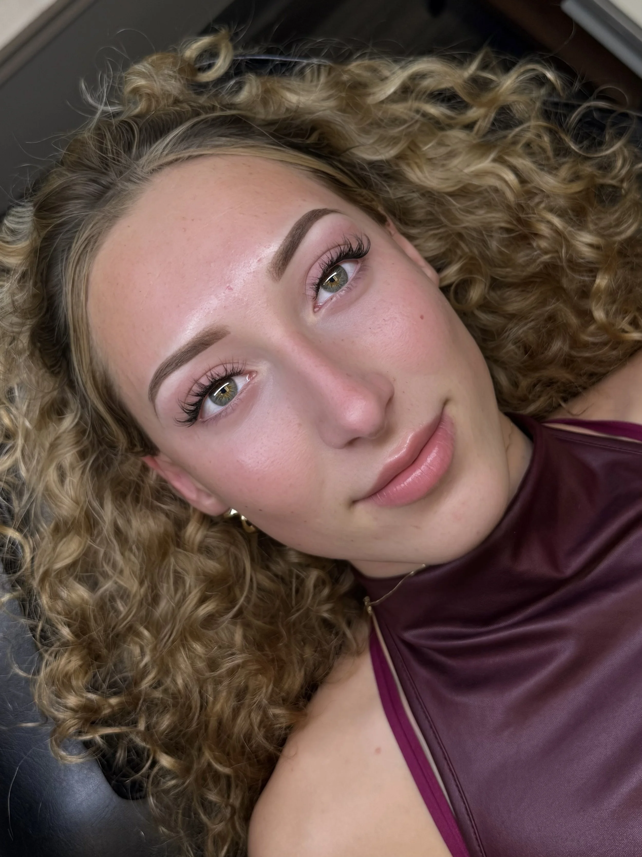 Close-up of a woman with curly blonde hair lying down, looking slightly to the side. She has light skin, green eyes, and is wearing makeup including eye shadow, mascara, and lipstick. She is dressed in a maroon sleeveless top with purple accents.