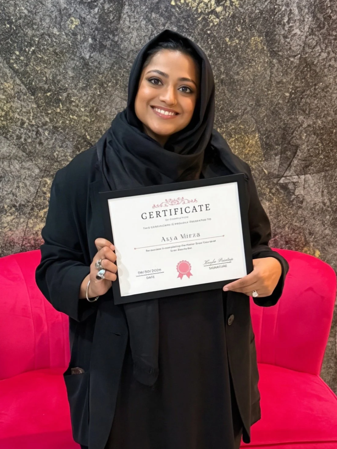 A woman wearing a black hijab and black coat holding a certificate, sitting on a pink sofa against a textured gray wall.