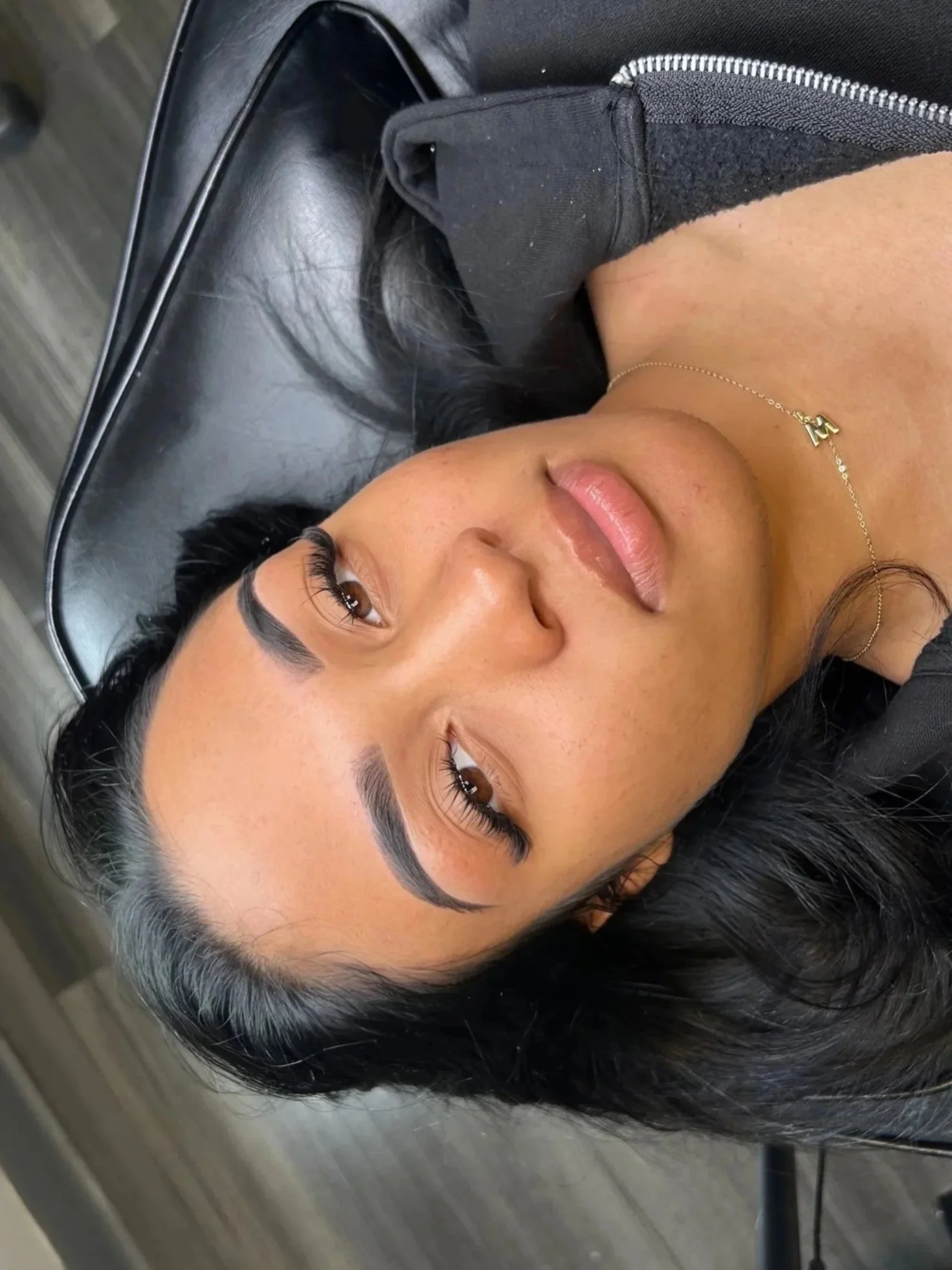 A woman with dark hair and tan skin lying on a black leather surface, wearing a black hoodie and a delicate gold necklace, with her eyes open.