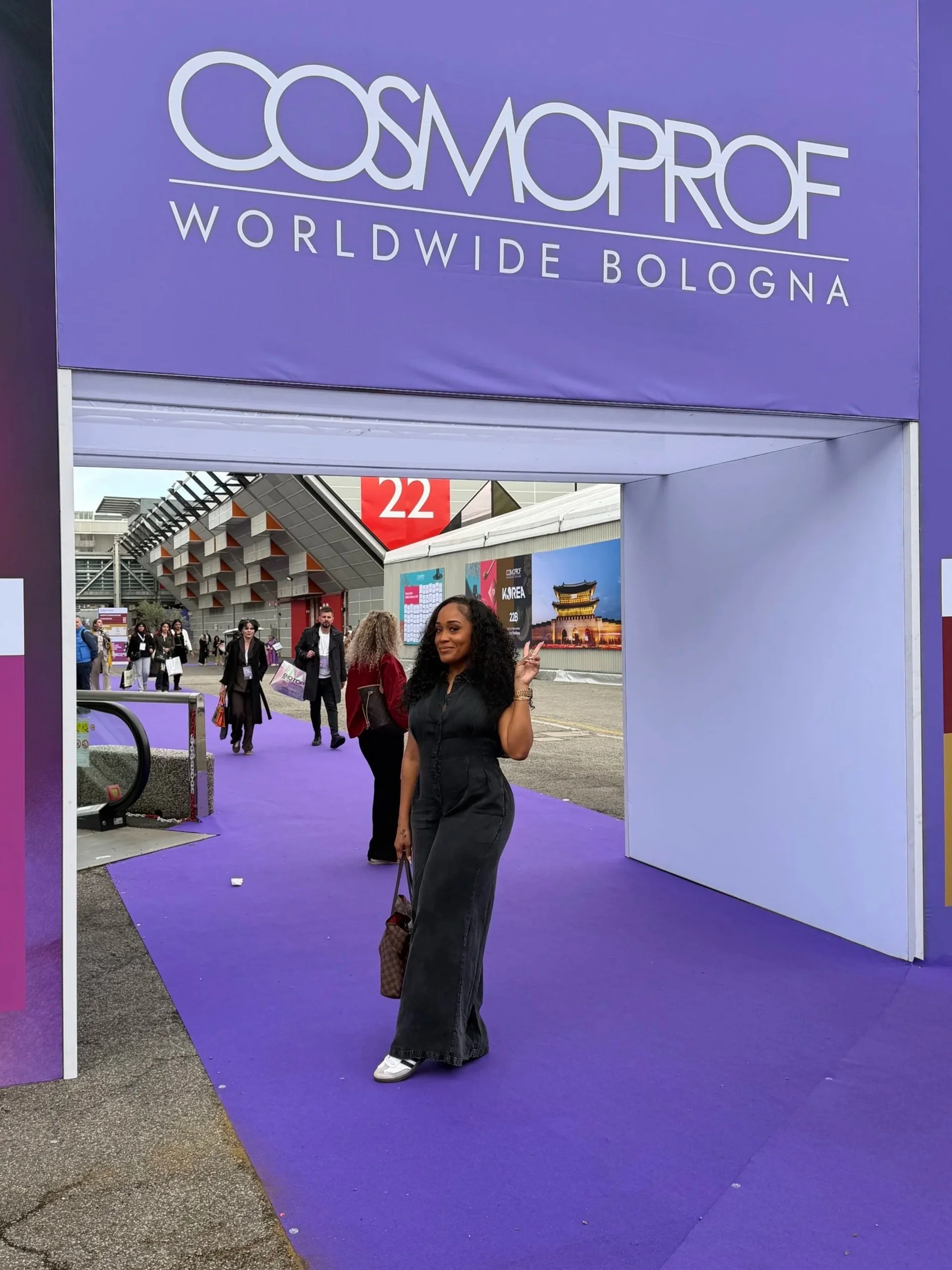 A woman standing on a purple carpet at the entrance of the Cosmoprof Worldwide Bologna event, pointing with a peace sign. Other attendees are walking in the background, and there are promotional banners visible.