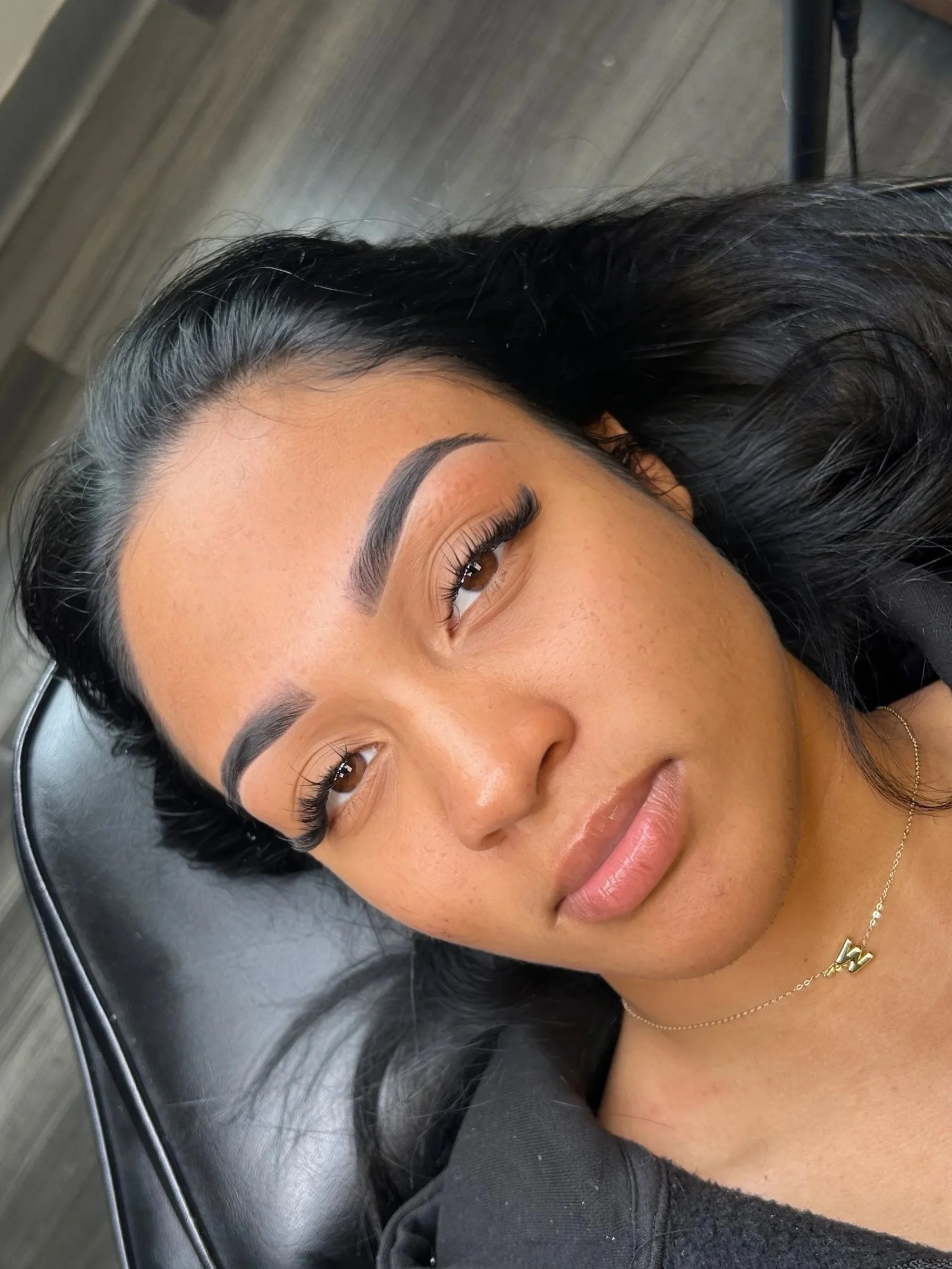 Close-up of a woman lying down on a black surface, showing her face with well-groomed eyebrows, long eyelashes, and black hair with some gray streaks. She is wearing a gold necklace and a black shirt.