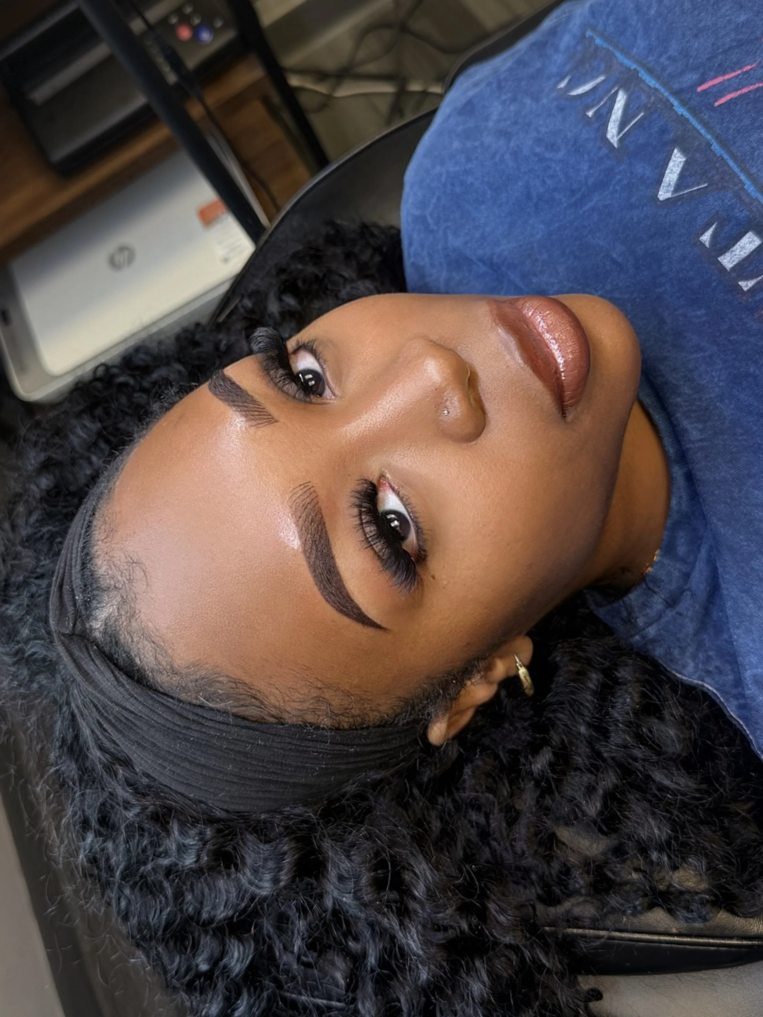 Close-up of a young woman with curly black hair, wearing a headband, with makeup including well-defined eyebrows, eye makeup, and glossy lips. She is lying down on a fluffy black and white blanket, wearing a blue shirt. In the background, there are electronic devices and cables.