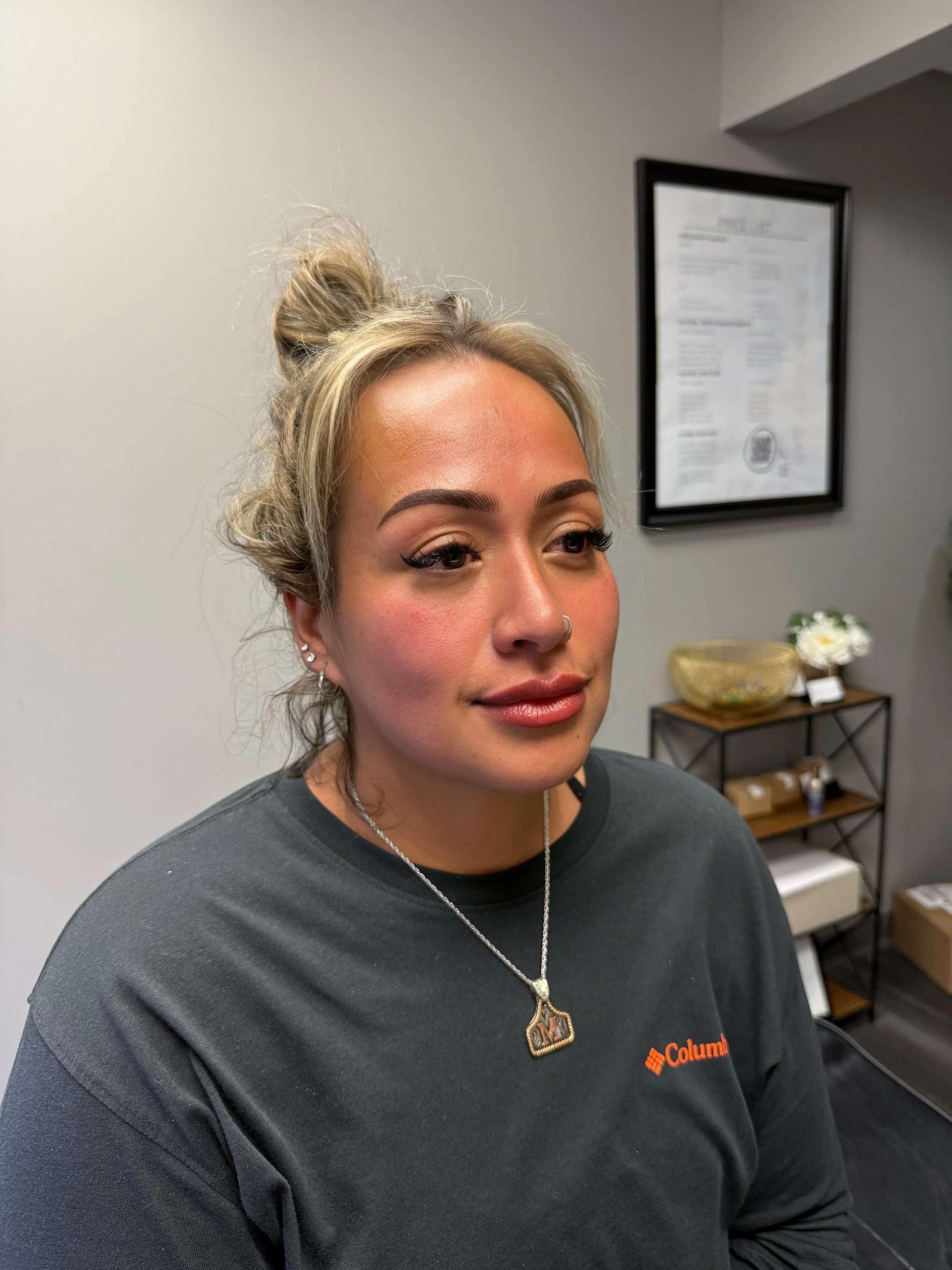 A woman with blonde hair styled in a messy bun, wearing makeup, a black Columbia sweatshirt, a silver necklace with a heart-shaped pendant, and earrings, sitting in an indoor space with a gray wall and framed picture in the background.