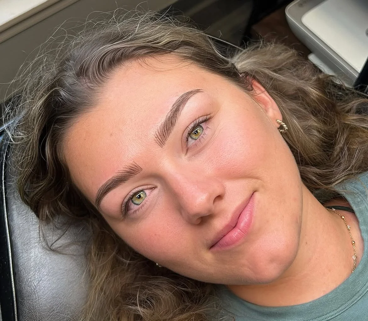 Close-up of a young woman with curly hair, lying on a medical chair or bed, smiling gently, wearing small earrings and a necklace, with natural makeup and clear skin.
