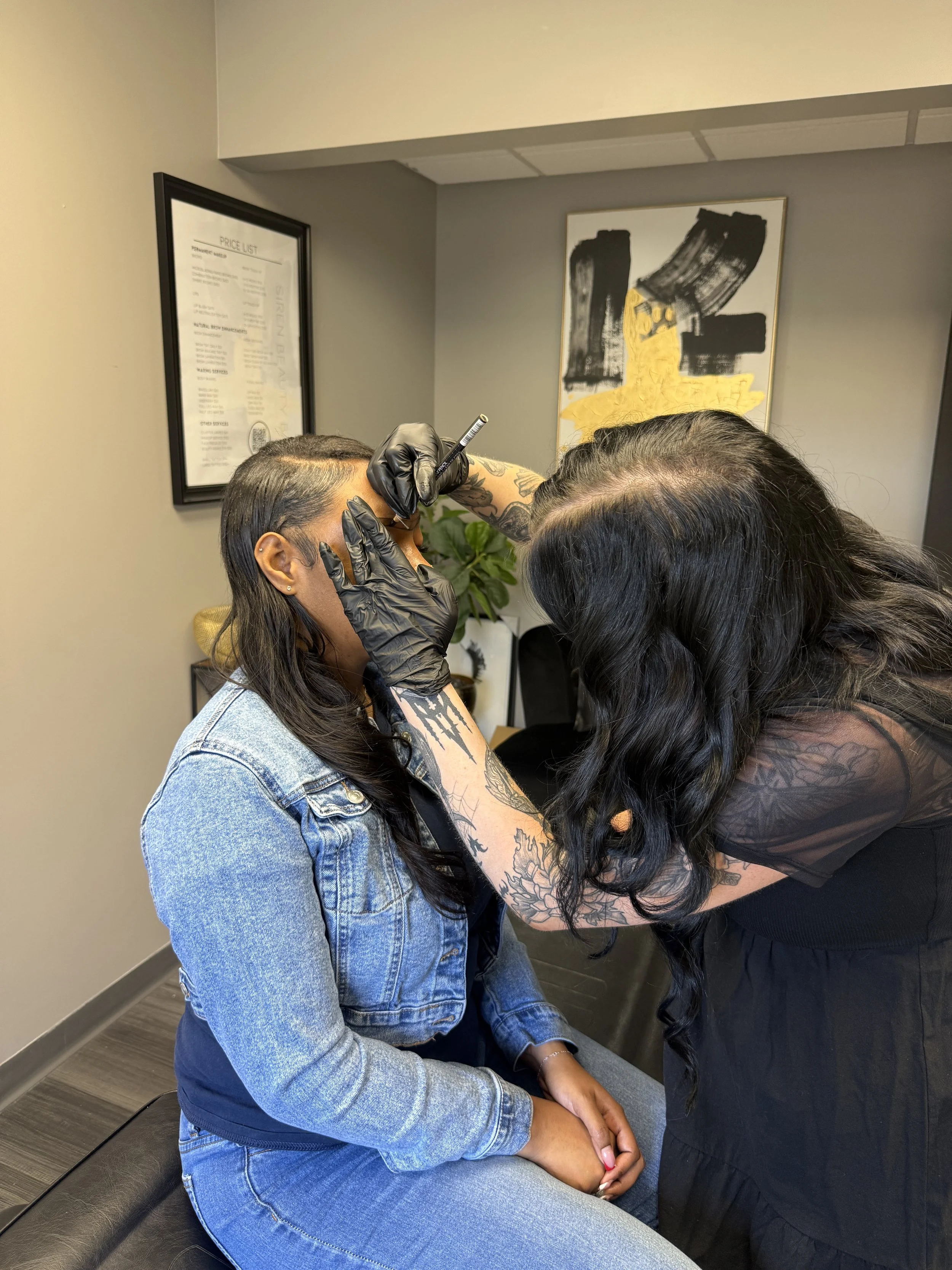 A person seated on a black bench in a tattoo studio, receiving a tattoo on her left arm from a tattoo artist wearing black gloves, with artwork and a price list on the walls behind them.