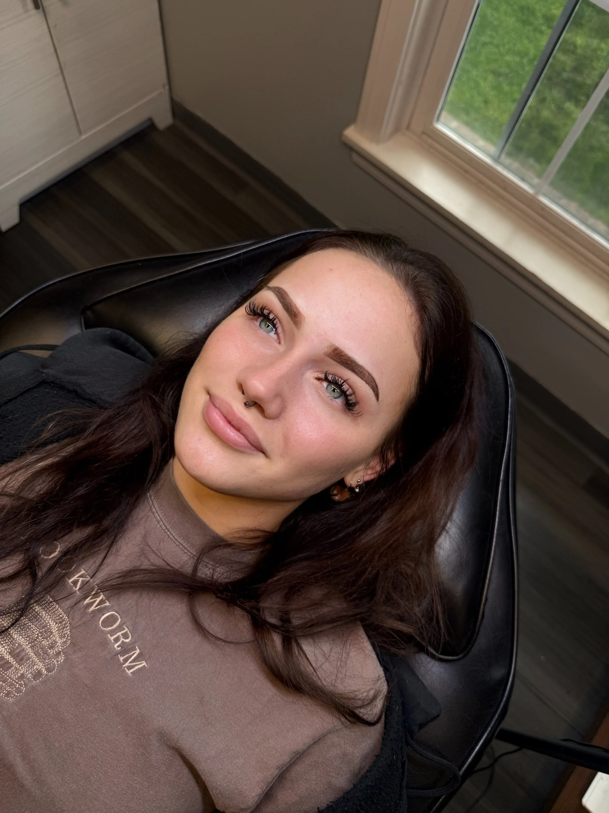 A young woman with dark hair, green eyes, and facial piercings, lying on a black chair near a window with green grass visible outside.