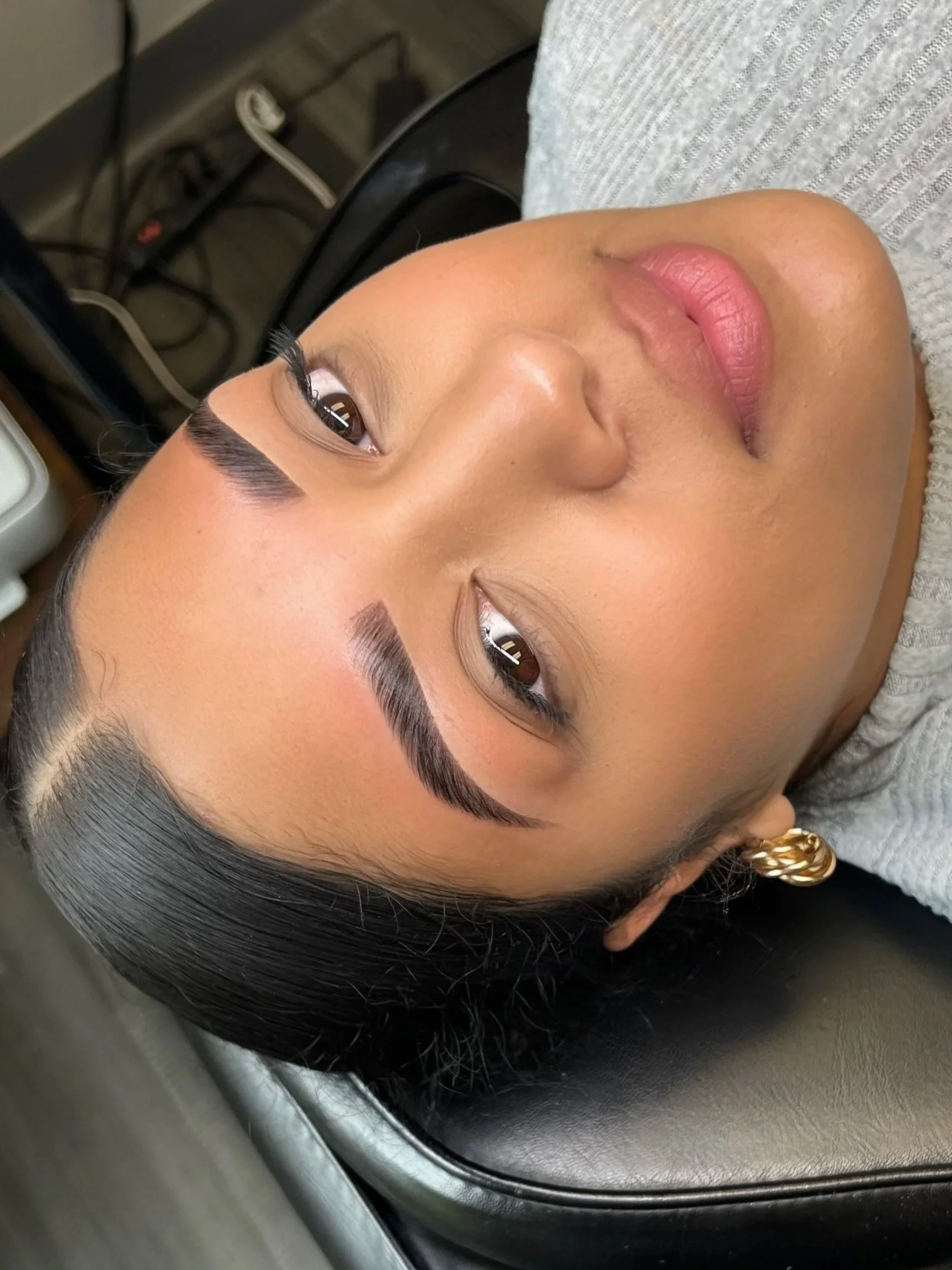 Close-up of a woman with dark hair, brown eyes, and gold hoop earrings lying down with her head on a gray chair or bed.