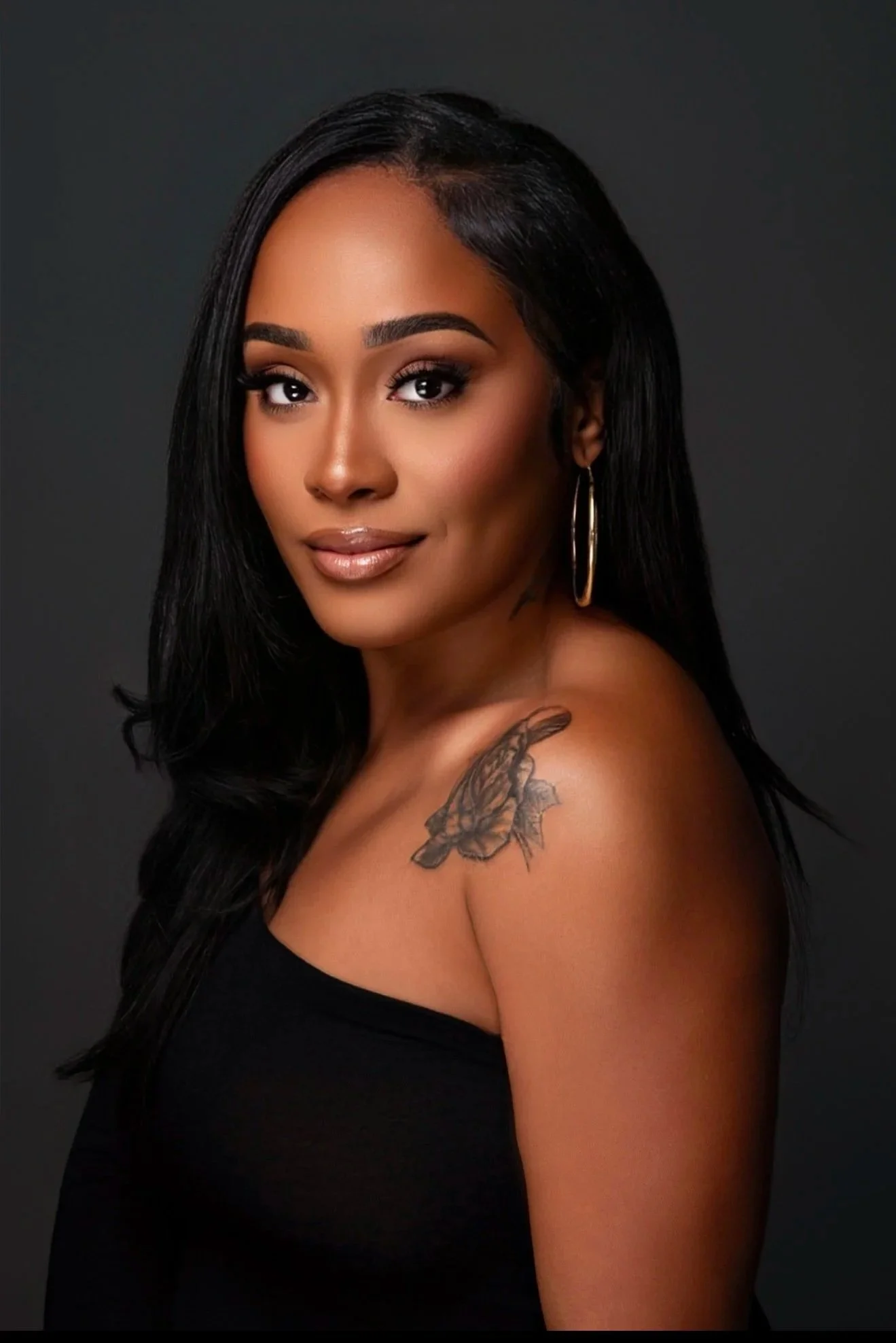 Portrait of a woman with long black hair, wearing a black strapless top, gold hoop earrings, and a butterfly tattoo on her shoulder, against a dark gray background.