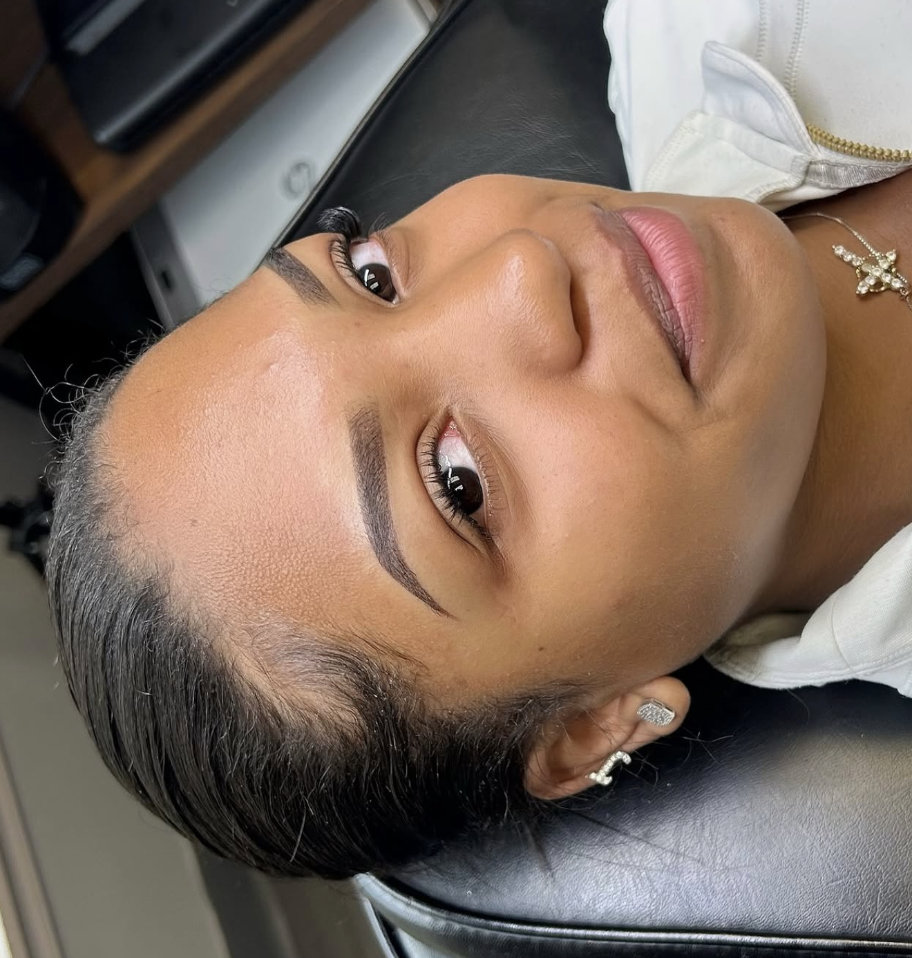 A woman with freshly applied eyebrow makeup, lying down with her head on a black surface, showing her face with a neutral expression, and wearing jewelry including earrings and a necklace.