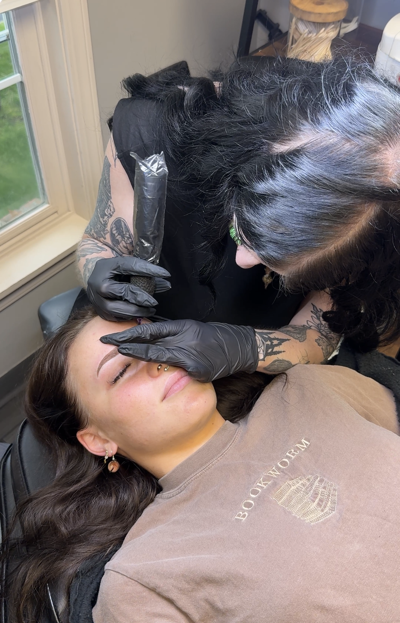 Tattooed woman receiving a tattoo on her forehead while lying down with eyes closed, tattoo artist wearing black gloves working on her forehead with tattoo equipment.