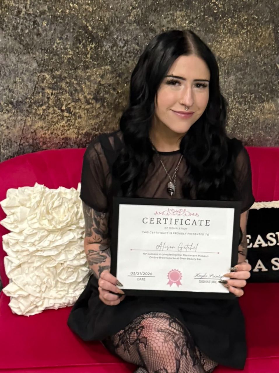 A woman with black hair and tattoos on her arms, holding a certificate, sitting on a red couch with textured wall background and white decorative pillow.
