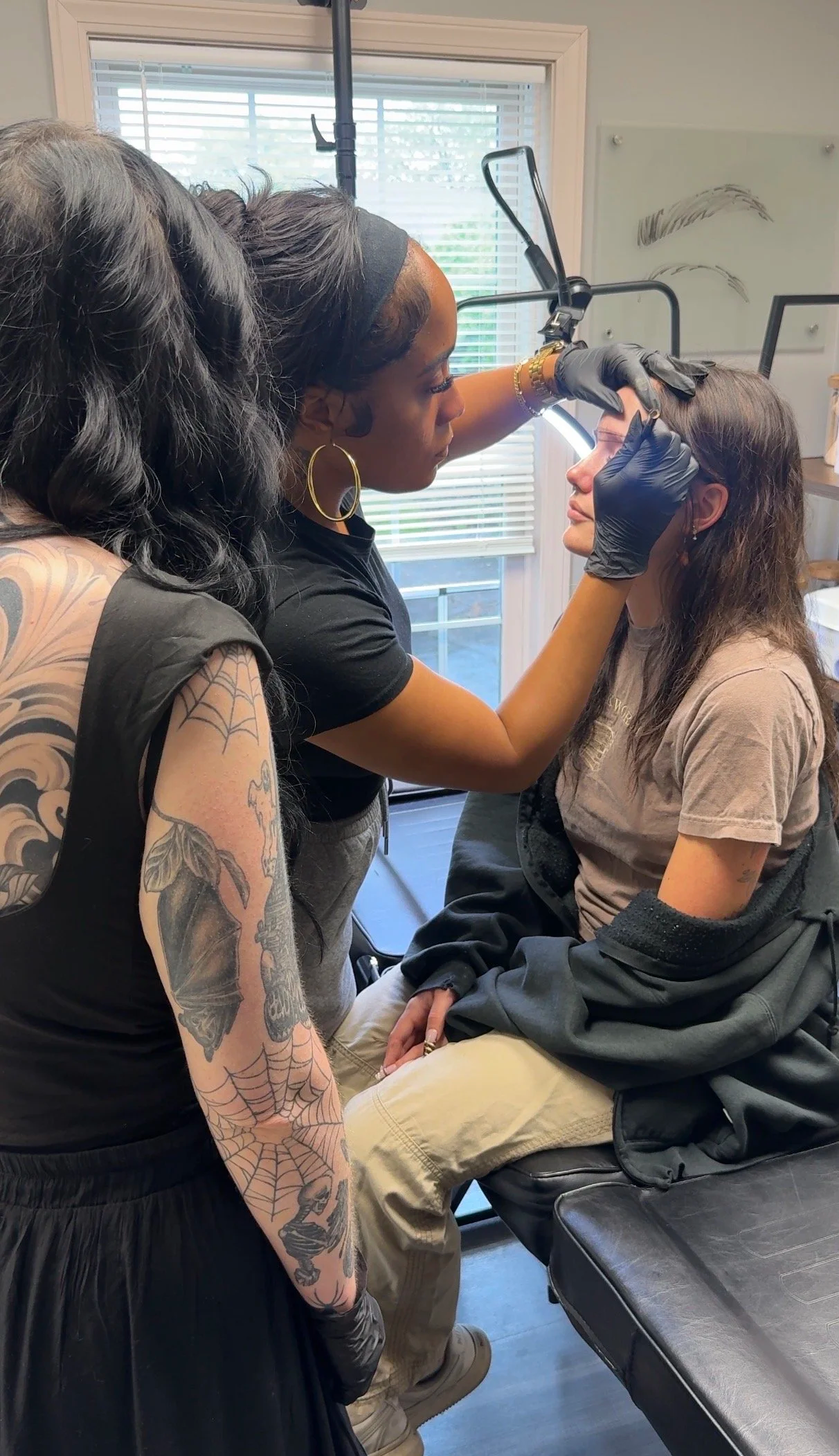 A tattooed woman with black hair and hoop earrings working on a girl with glasses, seated in a chair, in a tattoo or piercing studio. The woman is using a tool near the girl's face as she sits with her eyes closed. There is a window with blinds in the background.