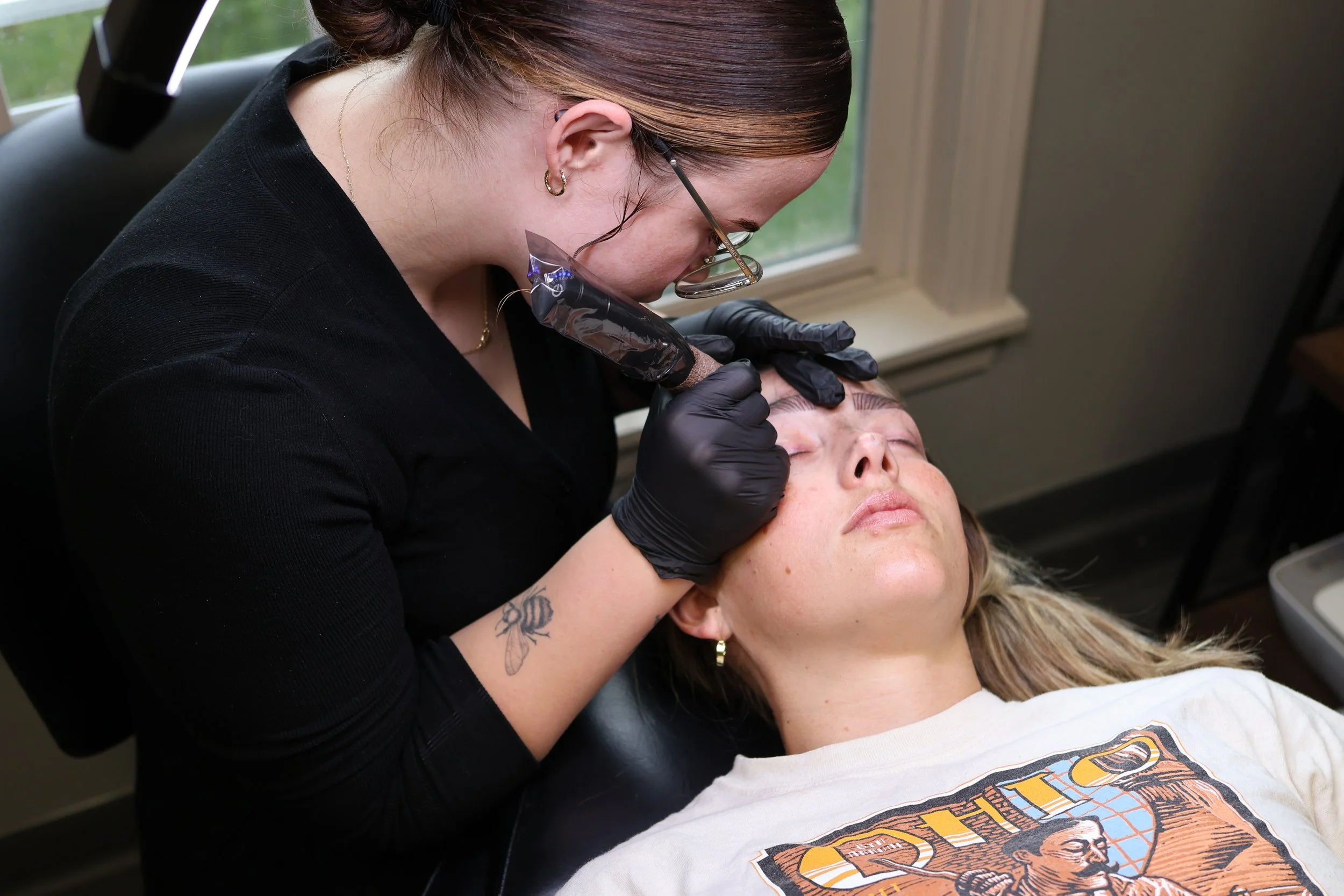 Tattooed woman with glasses tattooing a client's eyebrow in a salon.