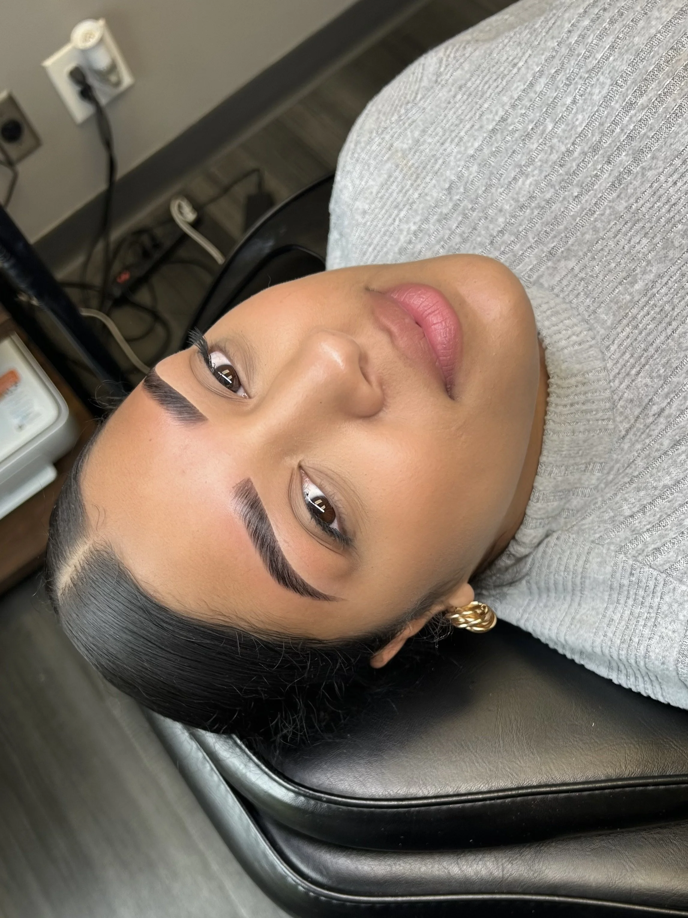 Close-up of a woman's face with makeup, lying on a black treatment table or bed, with a gray sweater and gold earrings, and dark hair styled straight.