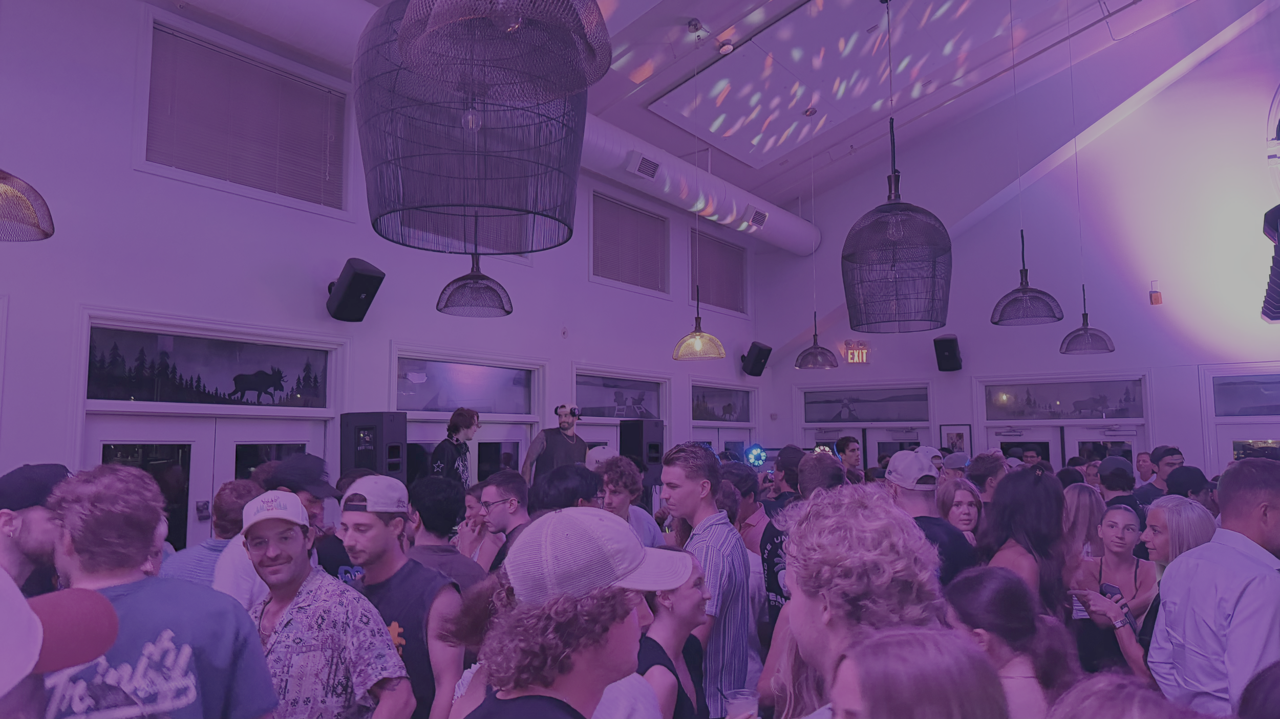 A crowded indoor party or concert with people socializing and dancing, decorated with hanging lamps and scenic window murals of nature.
