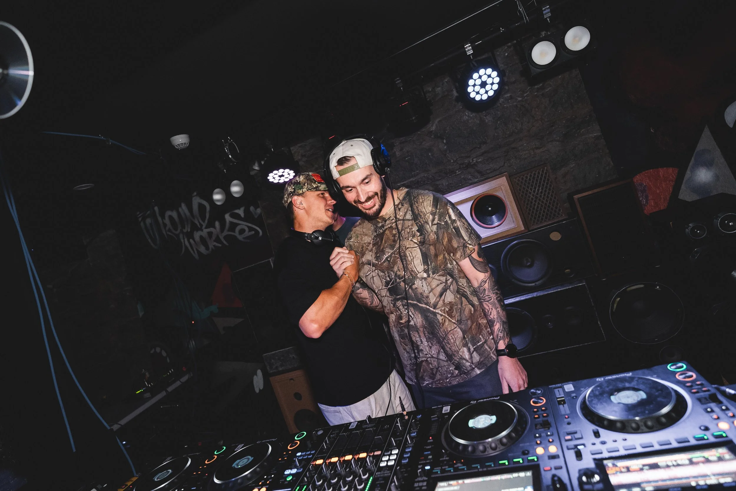 Two male DJs with headphones on, smiling and enjoying music in a dark club, surrounded by DJ equipment and speakers.