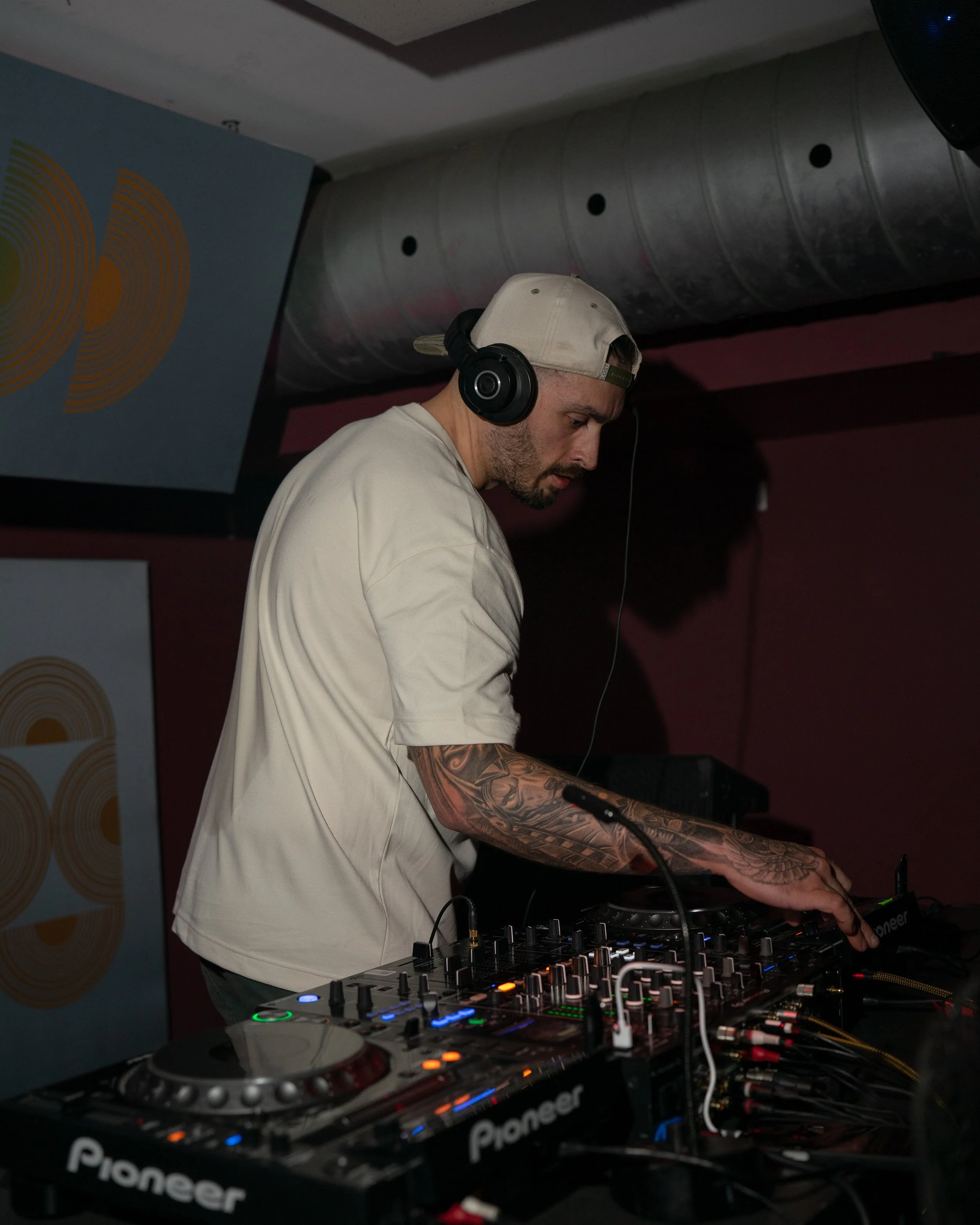 A DJ wearing a beige cap and headphones, focusing on mixing music with a Pioneer DJ controller, in a dimly lit venue.