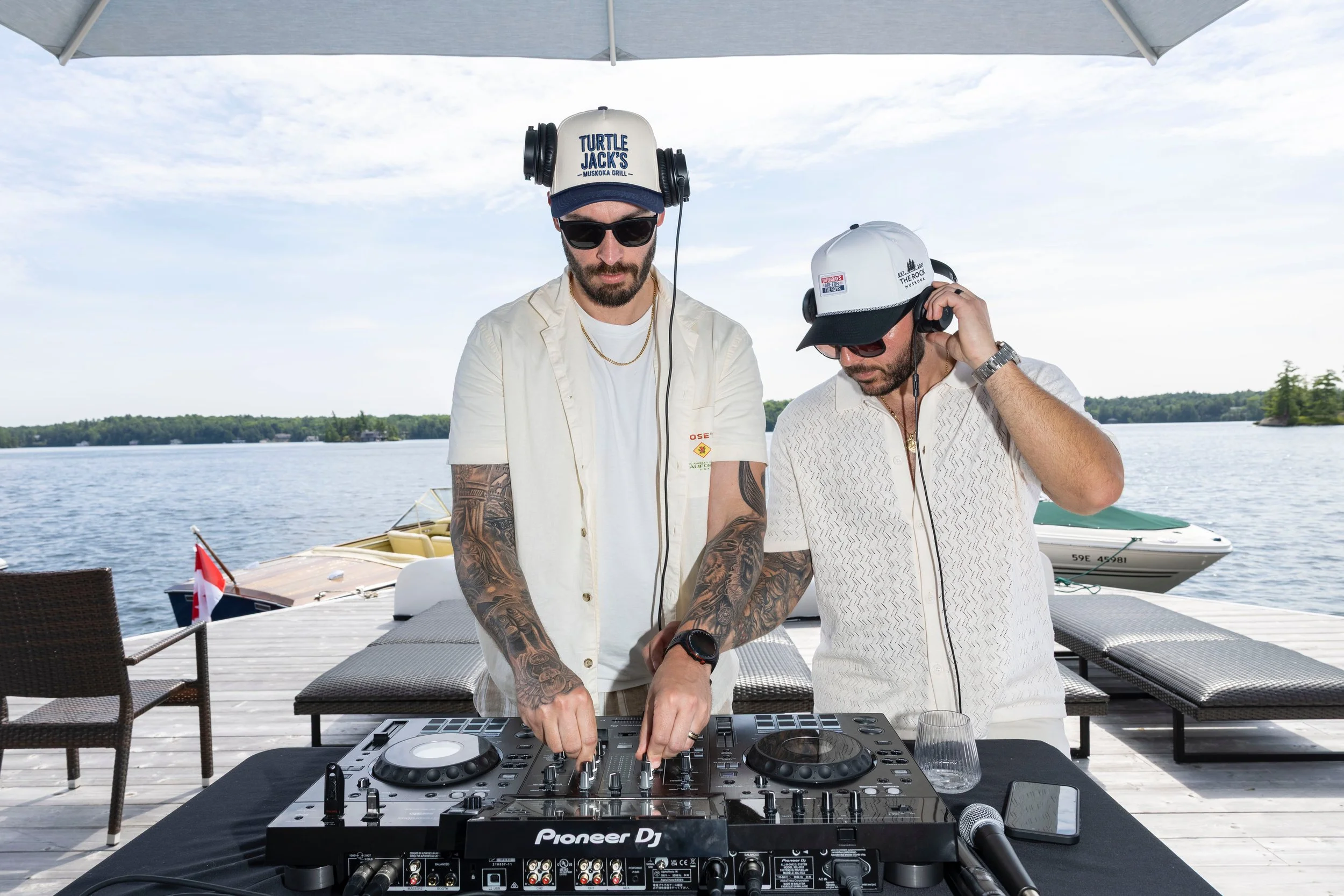 Two men are DJing outdoors on a deck by a body of water, with boats and trees in the background. One wears a white cap with a logo and headphones, the other wears a white hat and sunglasses. They are focused on a DJ controller labeled 'Pioneer DJ'.