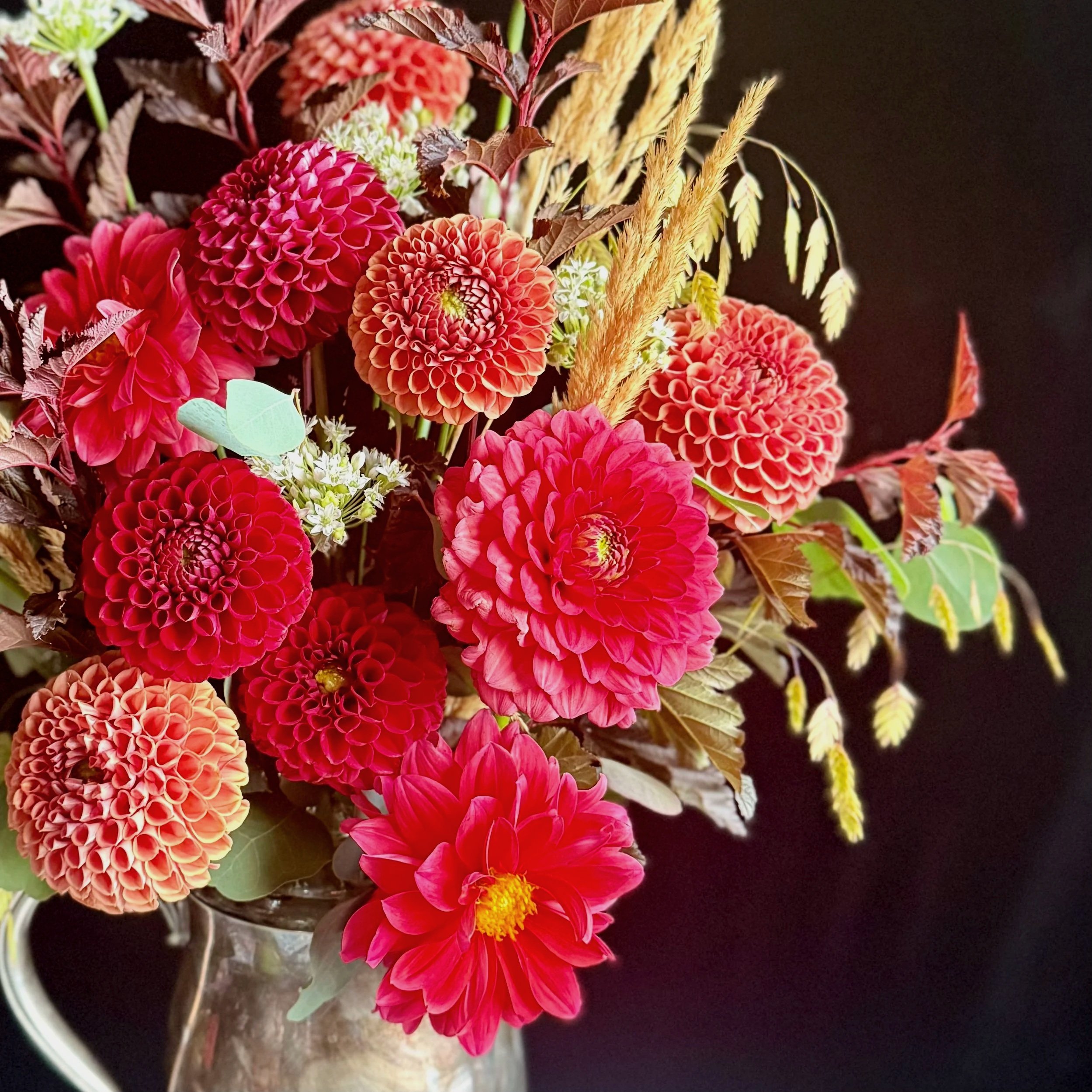 A vibrant floral arrangement in a glass vase featuring large red dahlias, pink flowers, small white blooms, yellowish-brown pampas grass, and various green and purple leaves on a dark background.