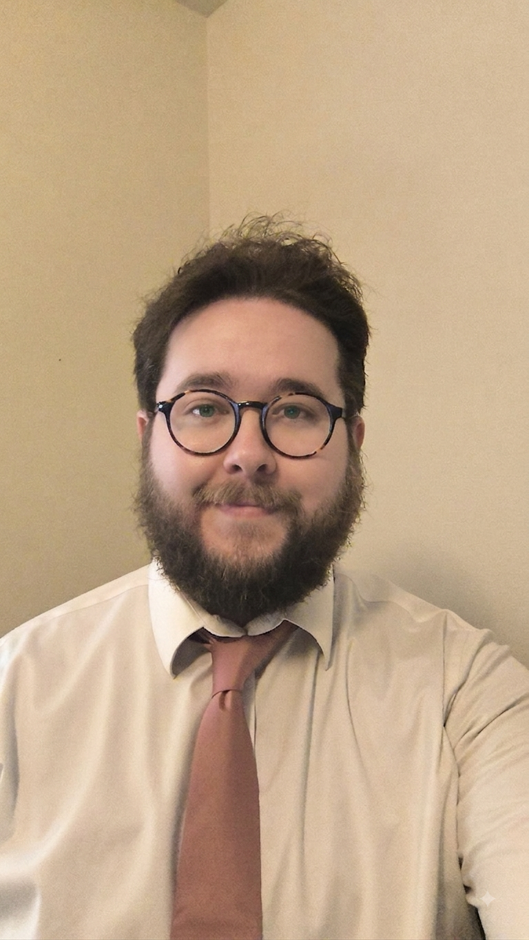A man with glasses, a beard, and curly hair taking a selfie against a beige wall, wearing a light-colored shirt and a pink tie.