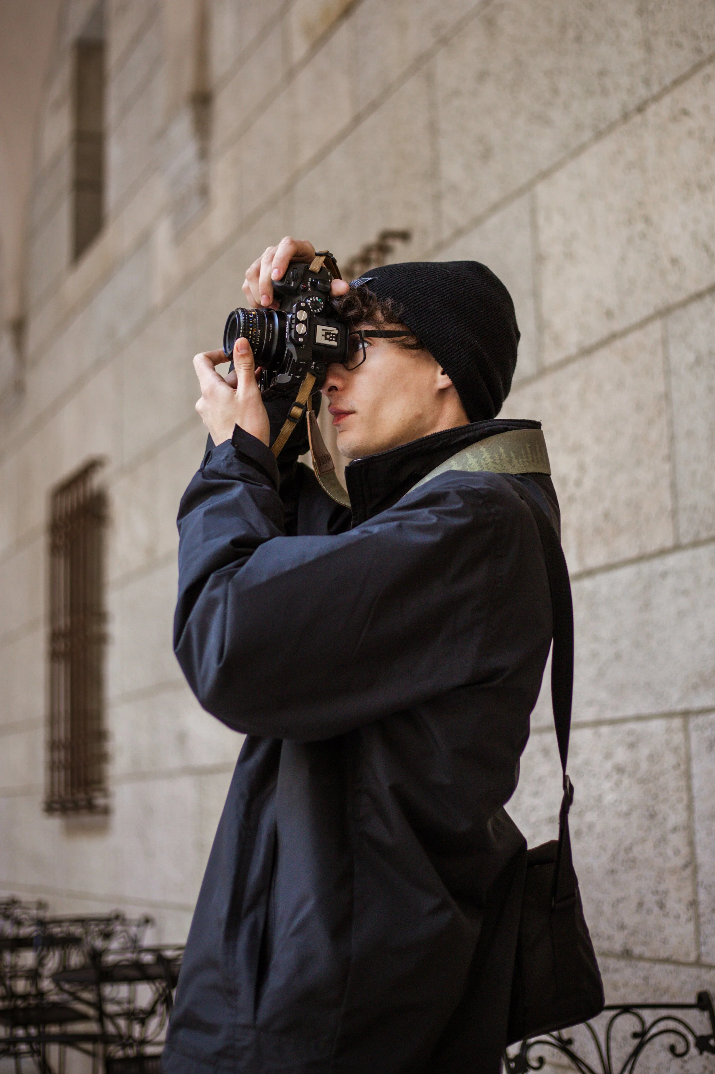 A person wearing a black beanie, glasses, and a black jacket, holding a camera up to their eye, taking a photo against a stone wall background.