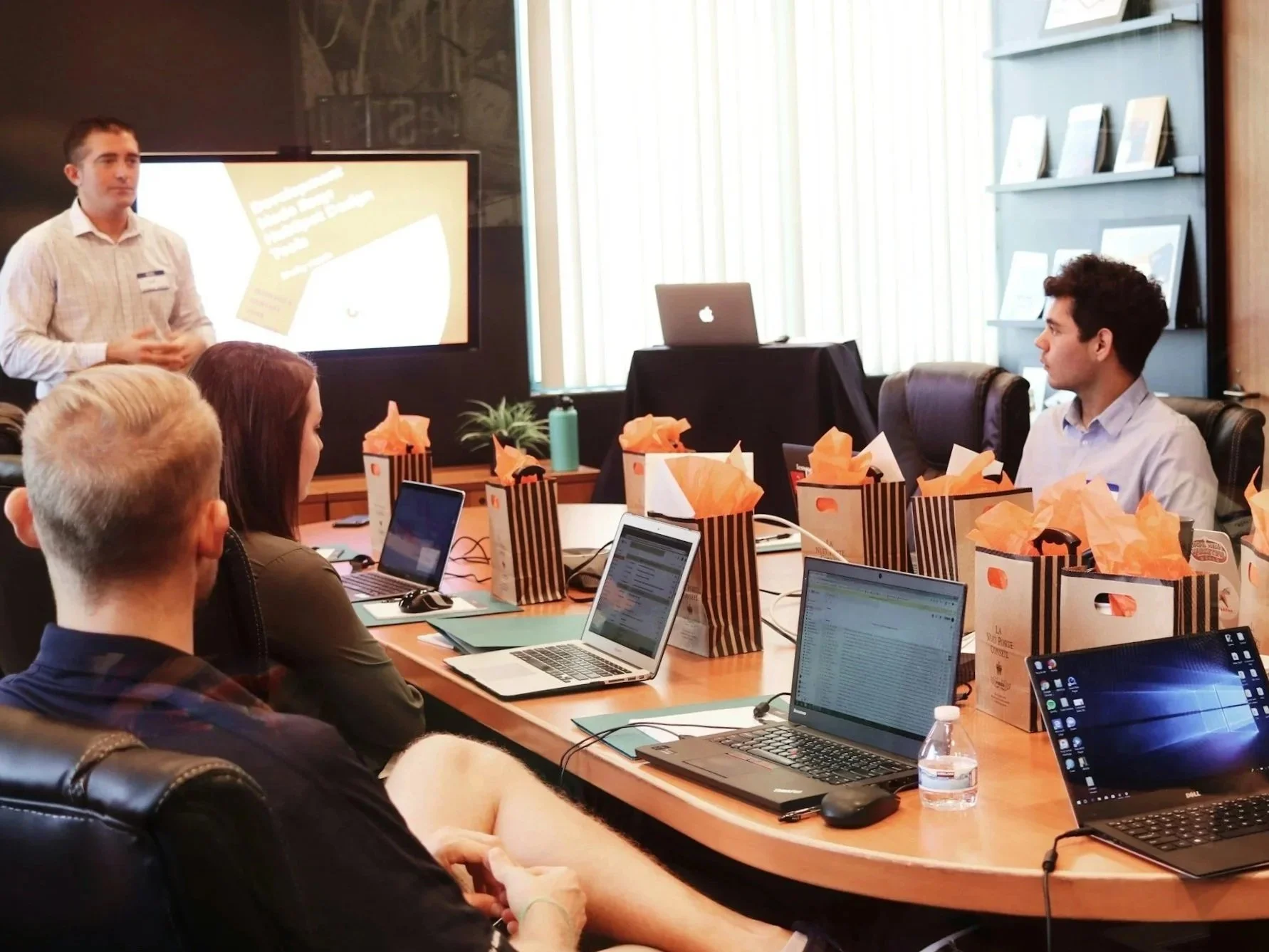 A group of people attending a meeting or workshop in a conference room, with a presenter standing near a large screen displaying a presentation, and laptops and gift bags on the table.
