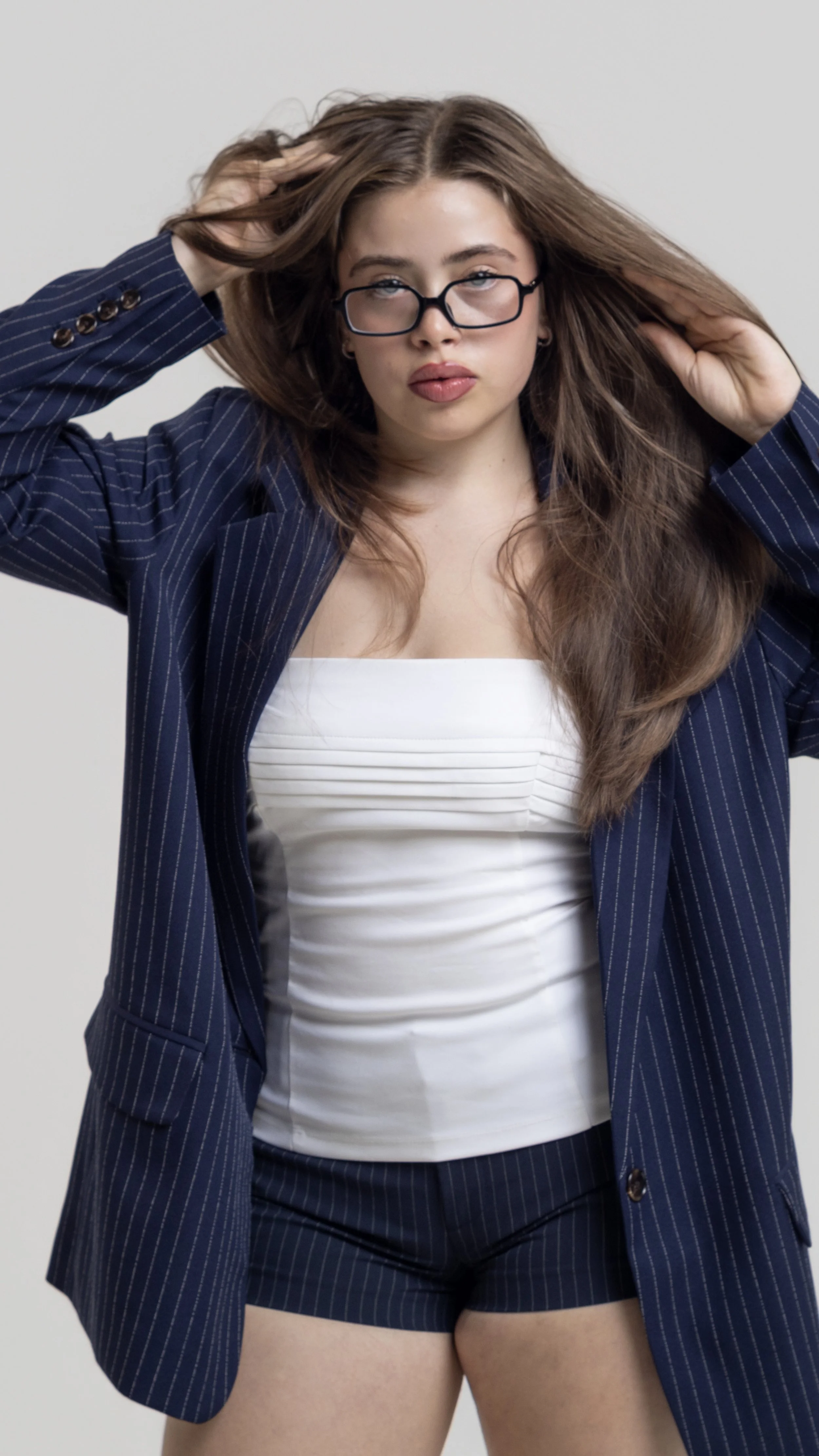 Young woman with long brown hair, glasses, and makeup, wearing a dark pinstripe blazer and matching shorts, with a white top, posing with her hands in her hair against a plain gray background.