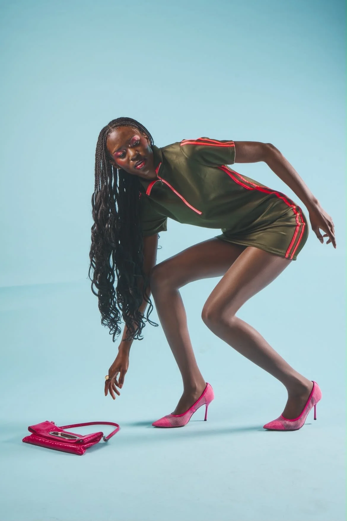 A woman with long, curly hair in a fashion pose wearing a shiny olive green dress with red stripes, pink high heels, and a matching pink purse placed on the floor.