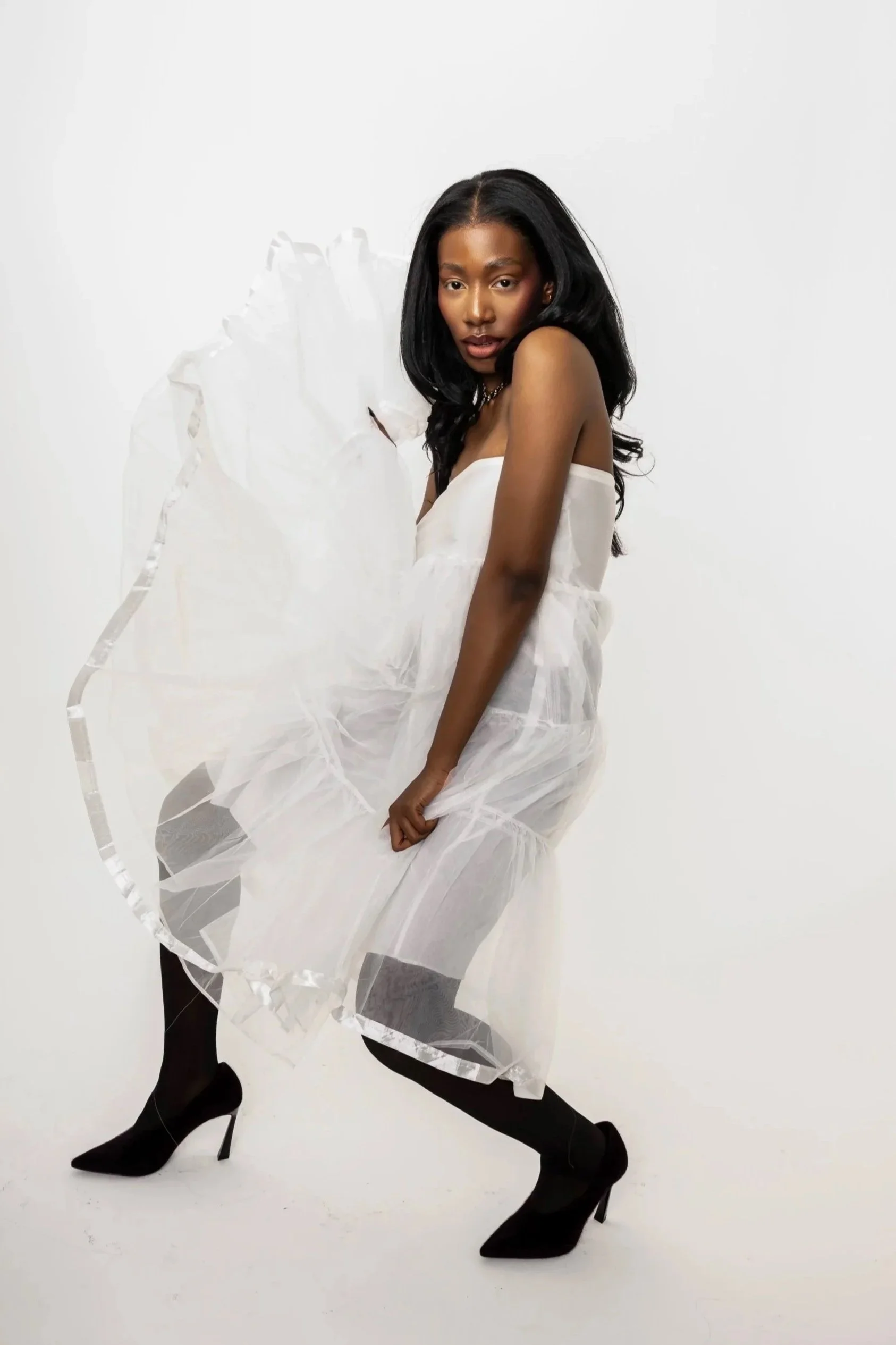 A woman with long black hair, wearing a strapless white dress, black tights, and black high heels, poses against a plain white background, holding a large transparent circular acrylic art piece.