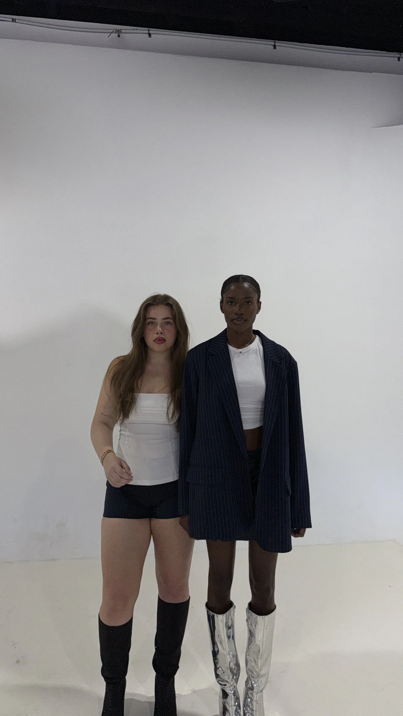 Two women standing in front of a white backdrop, dressed in fashionable outfits, with the woman on the left wearing a white tank top, shorts, and black knee-high socks, and the woman on the right wearing a pinstripe blazer and reflective silver boots.