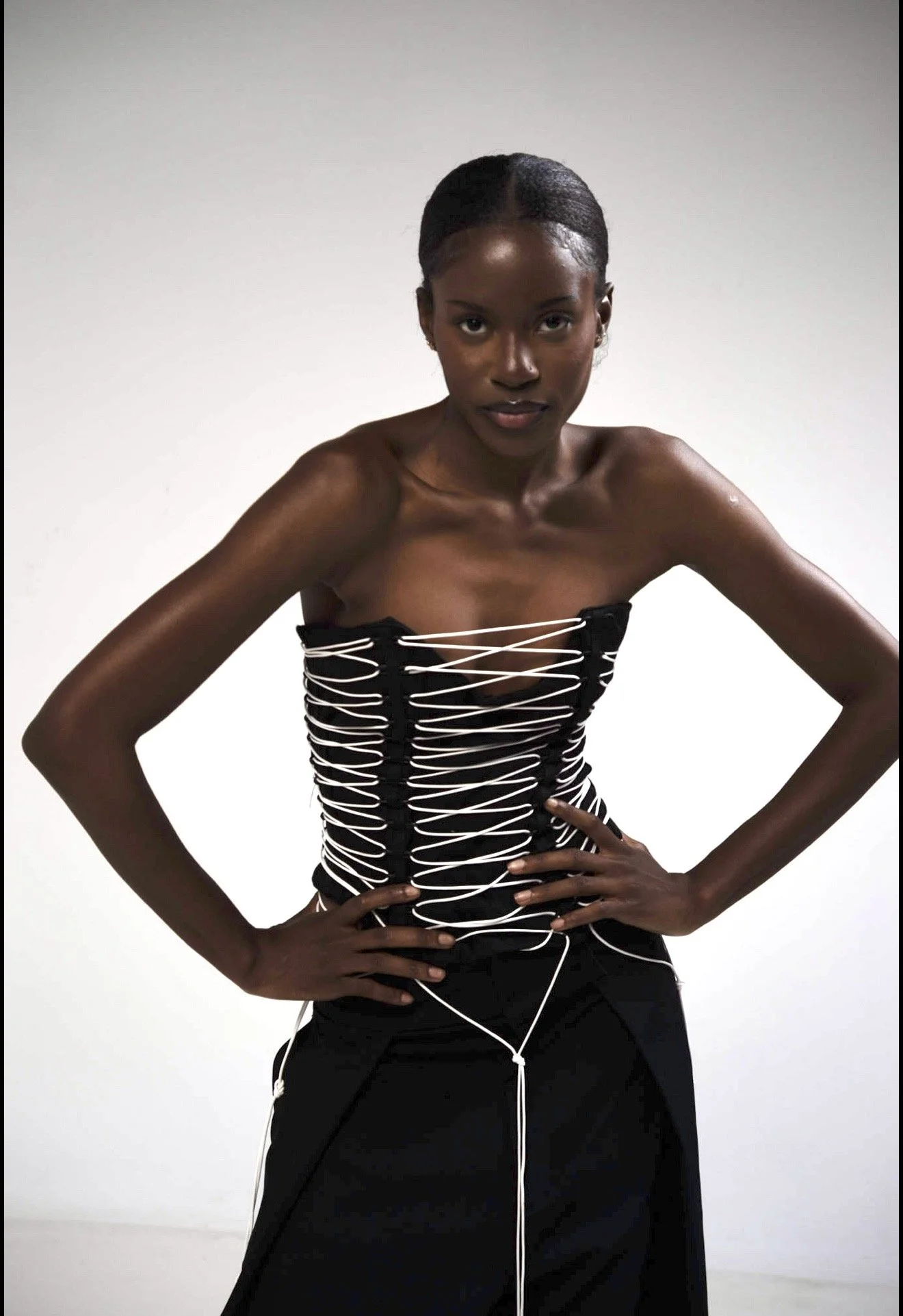 A woman with dark skin and short hair, wearing a black corset with white laces and a long black skirt, standing against a plain white background with her hands on her hips.