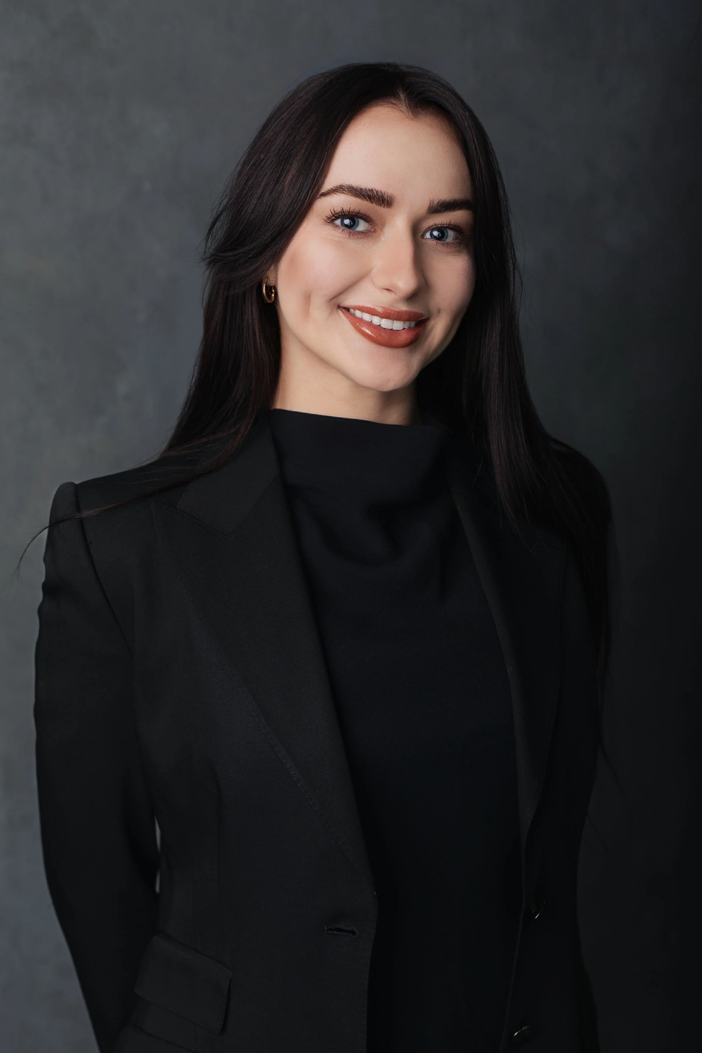 A woman with long dark hair, blue eyes, and light skin, dressed in a black blazer and black turtleneck, smiling against a dark gray background.
