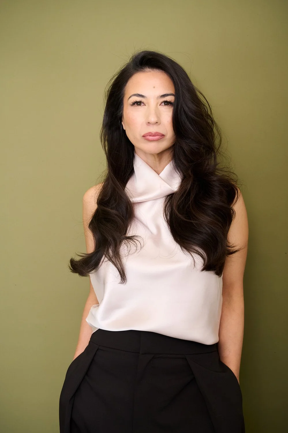 A woman with long, dark, wavy hair, wearing a white sleeveless top and black pants, standing against a light green background.