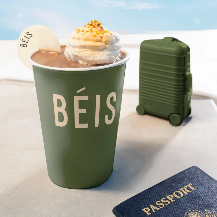 Green BÉIS branded hot chocolate cup beside a matching suitcase and passport